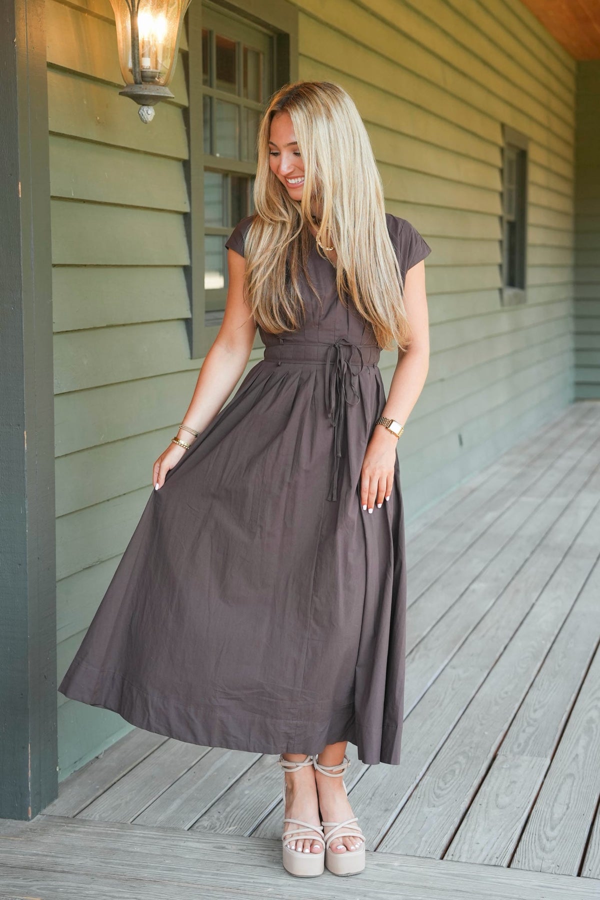 A woman stands on a wooden porch wearing a dark brown midi dress with short sleeves, a V-neckline, and a self-tie waist. She is wearing beige platform sandals and standing in front of a house with green siding.