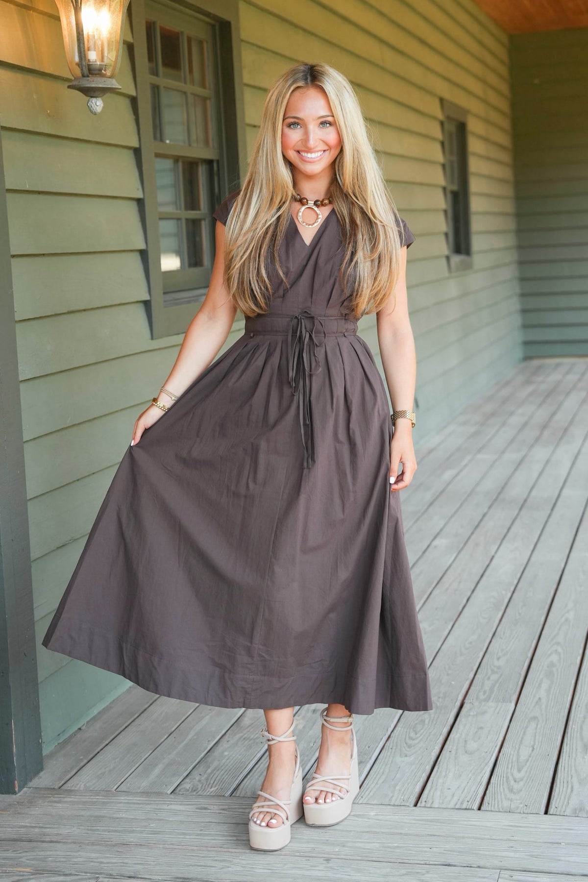 A woman stands on a wooden porch wearing a dark brown midi dress with short sleeves, a V-neckline, and a self-tie waist. She is wearing beige platform sandals and standing in front of a house with green siding.