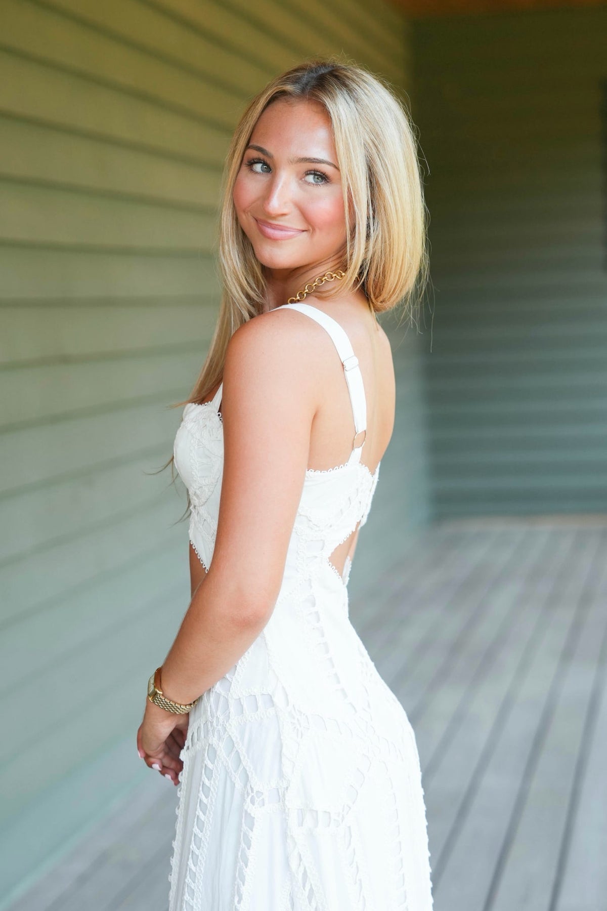 A woman stands on a wooden porch wearing a white sleeveless maxi dress with a sweetheart neckline, a midriff cutout, and an allover eyelet lace pattern.