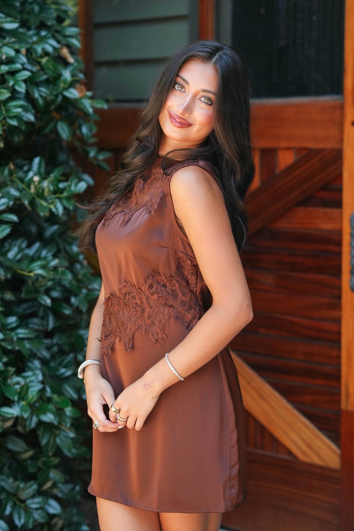 A woman wearing a sleeveless brown dress with lace detailing on the bodice and waist, standing outdoors in front of a wooden door and green foliage.