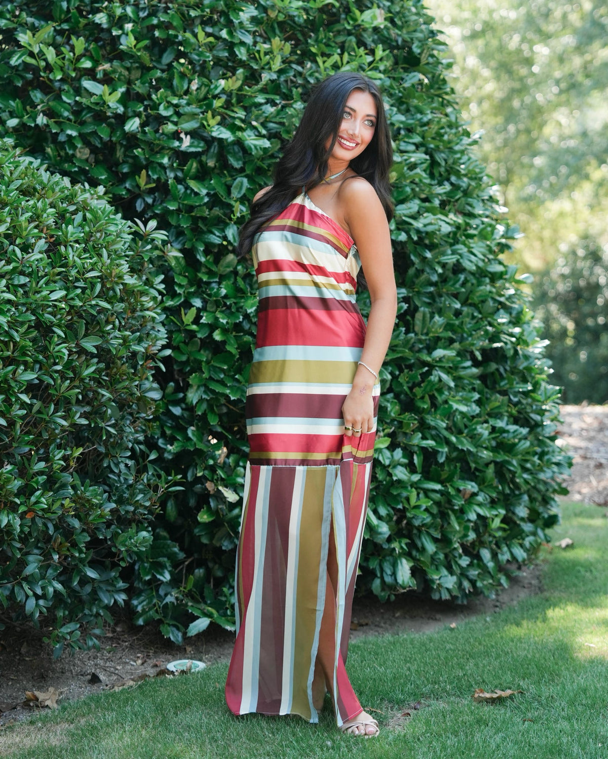 A woman wearing a colorful striped halter maxi dress stands outdoors in front of a wooden door and green foliage. The dress features vertical stripes in red, orange, yellow, green, and white.
