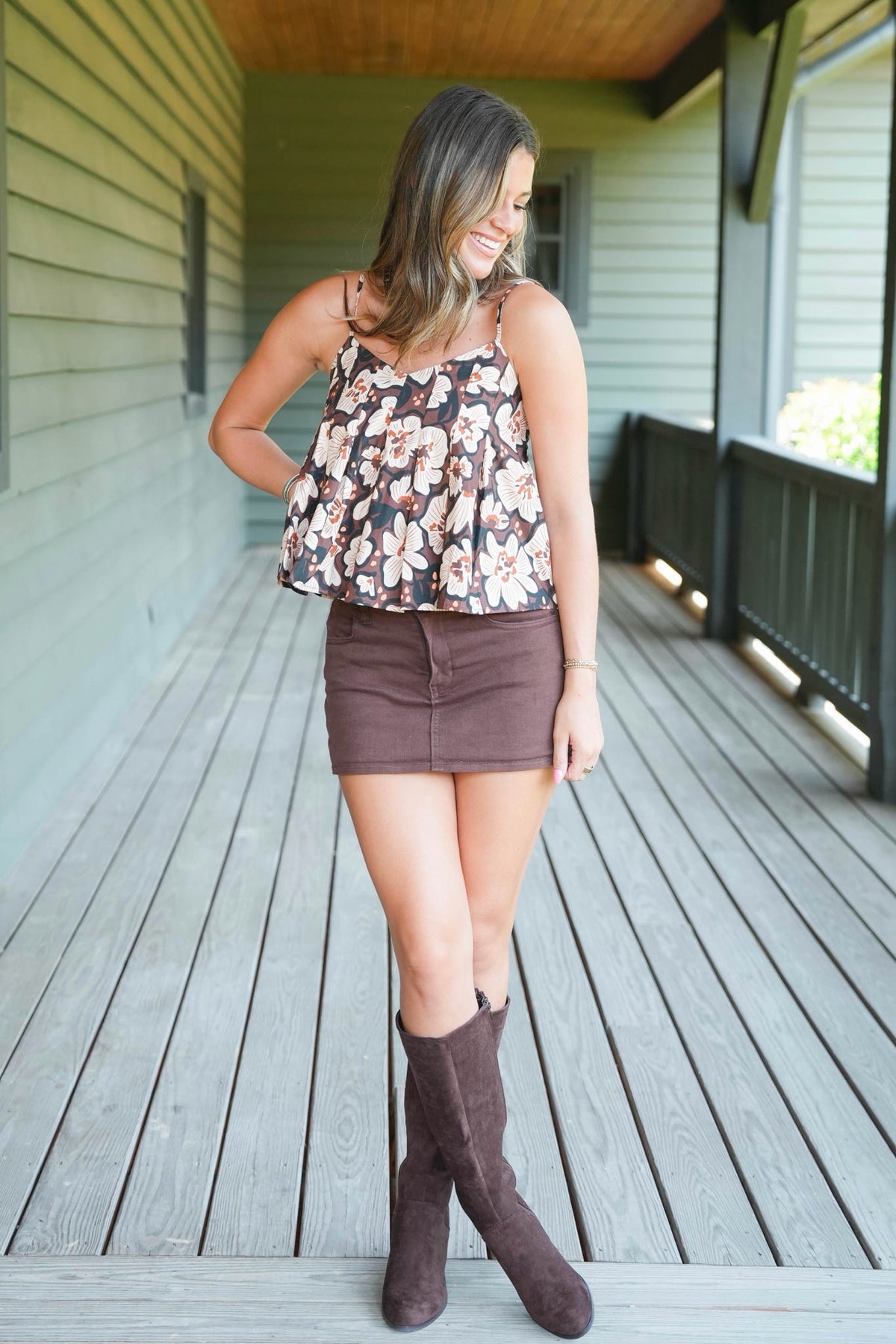 A woman stands on a wooden porch wearing a floral spaghetti-strap top, a brown corduroy mini skirt, and brown suede boots.