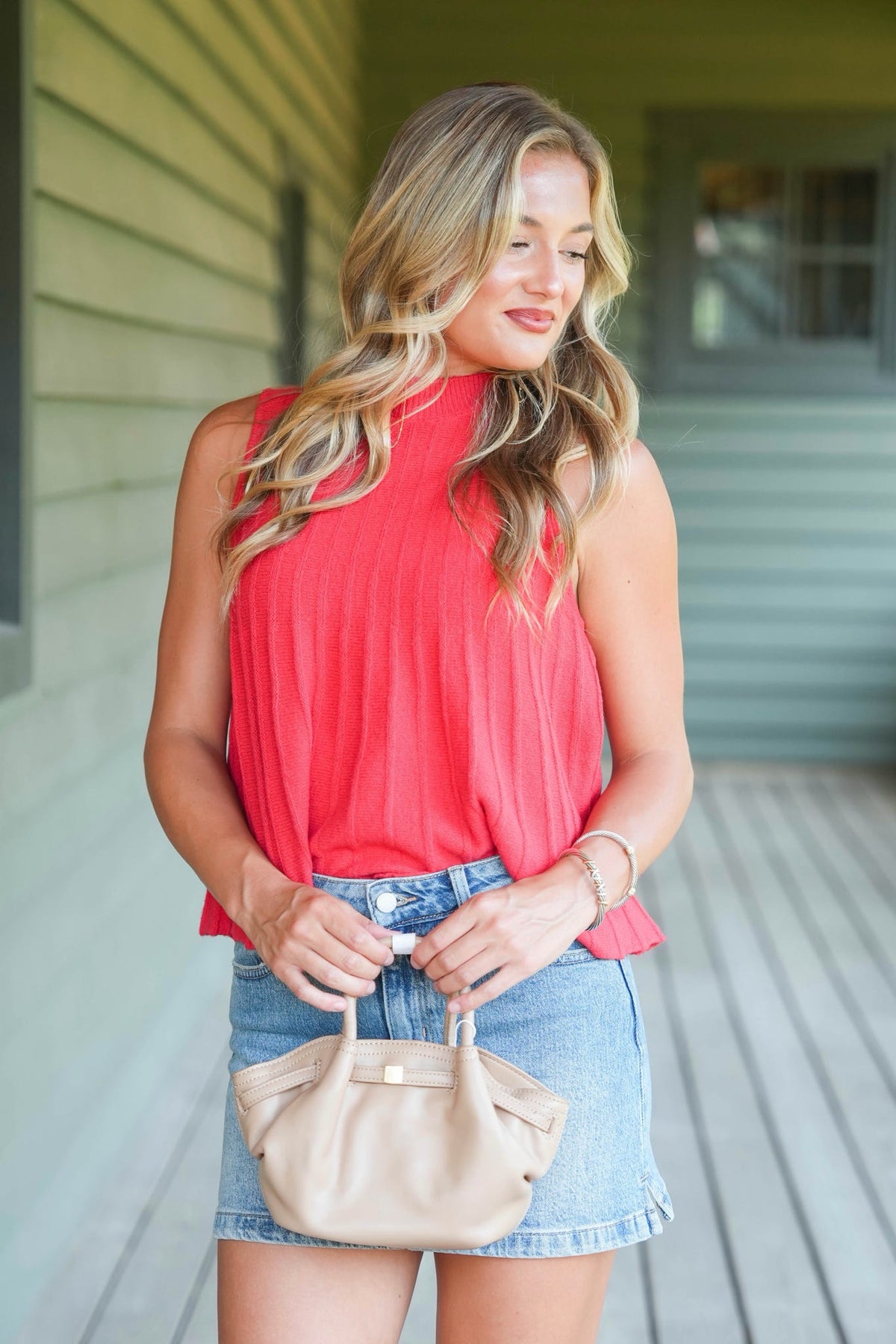 A woman stands on a porch wearing a sleeveless coral-red top and denim shorts, holding a beige handbag.