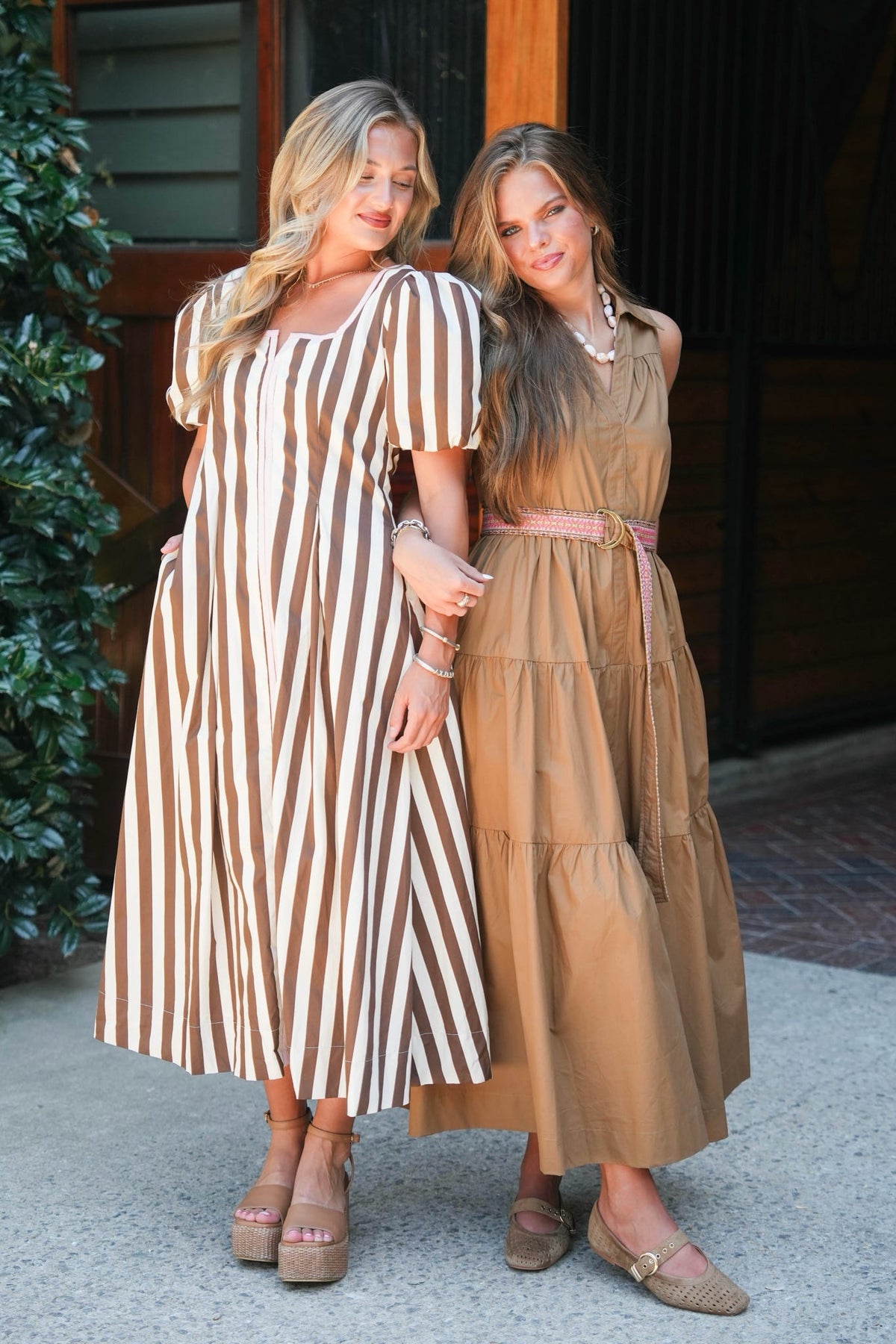 Two women standing side-by-side in tiered midi dresses. The woman on the left wears a brown and cream vertical striped dress with short puff sleeves.