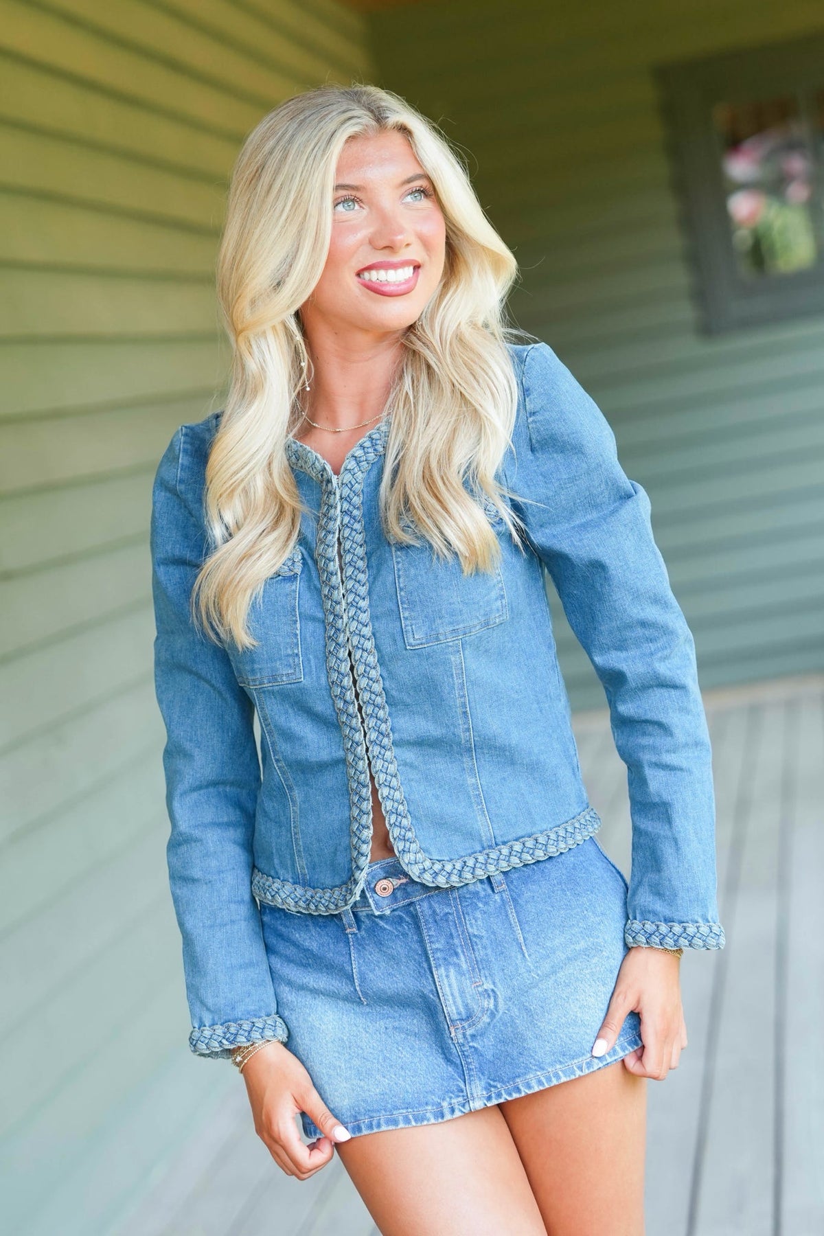 A smiling blonde woman wearing a light blue denim jacket and matching denim mini skirt with decorative braided trim, standing outdoors against a green house siding background.