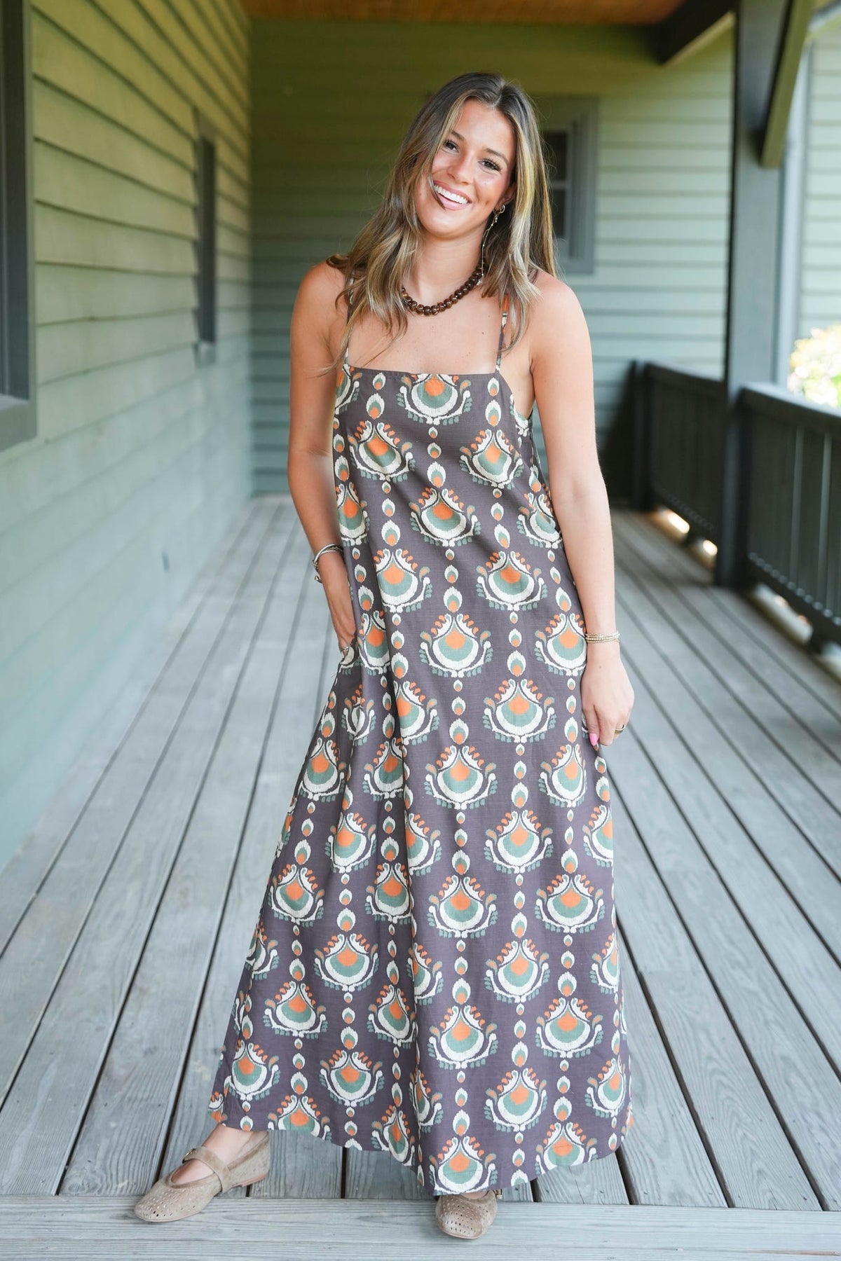 A woman wearing a sleeveless maxi dress with a brown, orange, and teal bohemian-style pattern, standing on a wooden deck in front of a house with green siding.