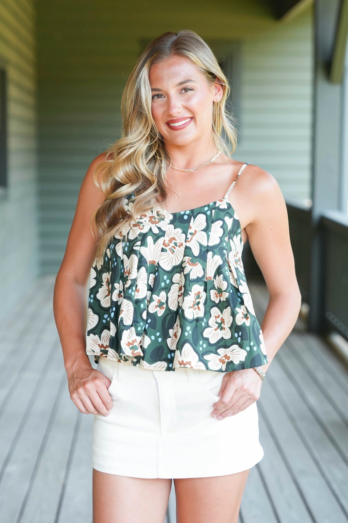 A woman modeling a dark green floral camisole top with white and beige flowers, paired with white shorts, standing on a wooden porch.