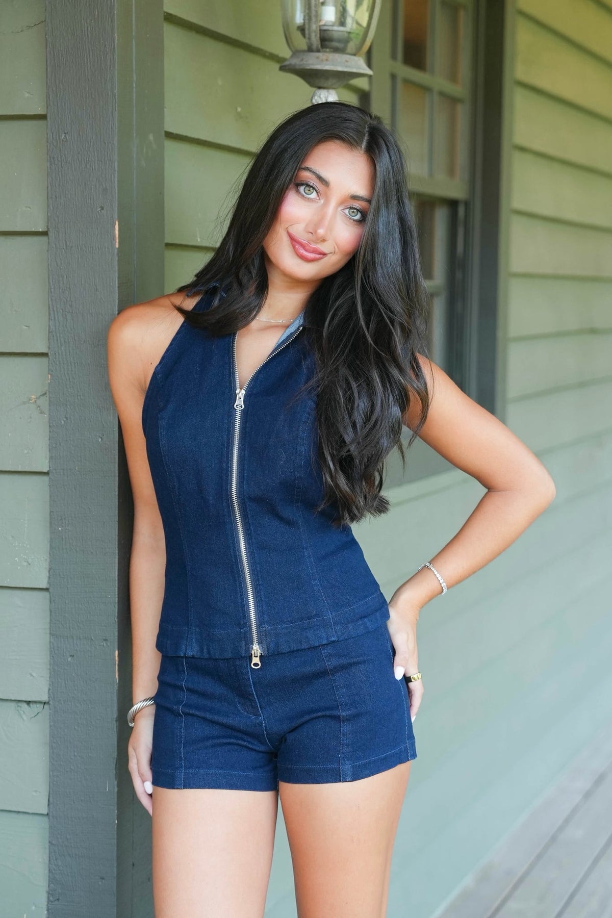 A woman modeling a dark blue denim halter top and matching denim shorts with a silver zipper, posing in front of a green house exterior.