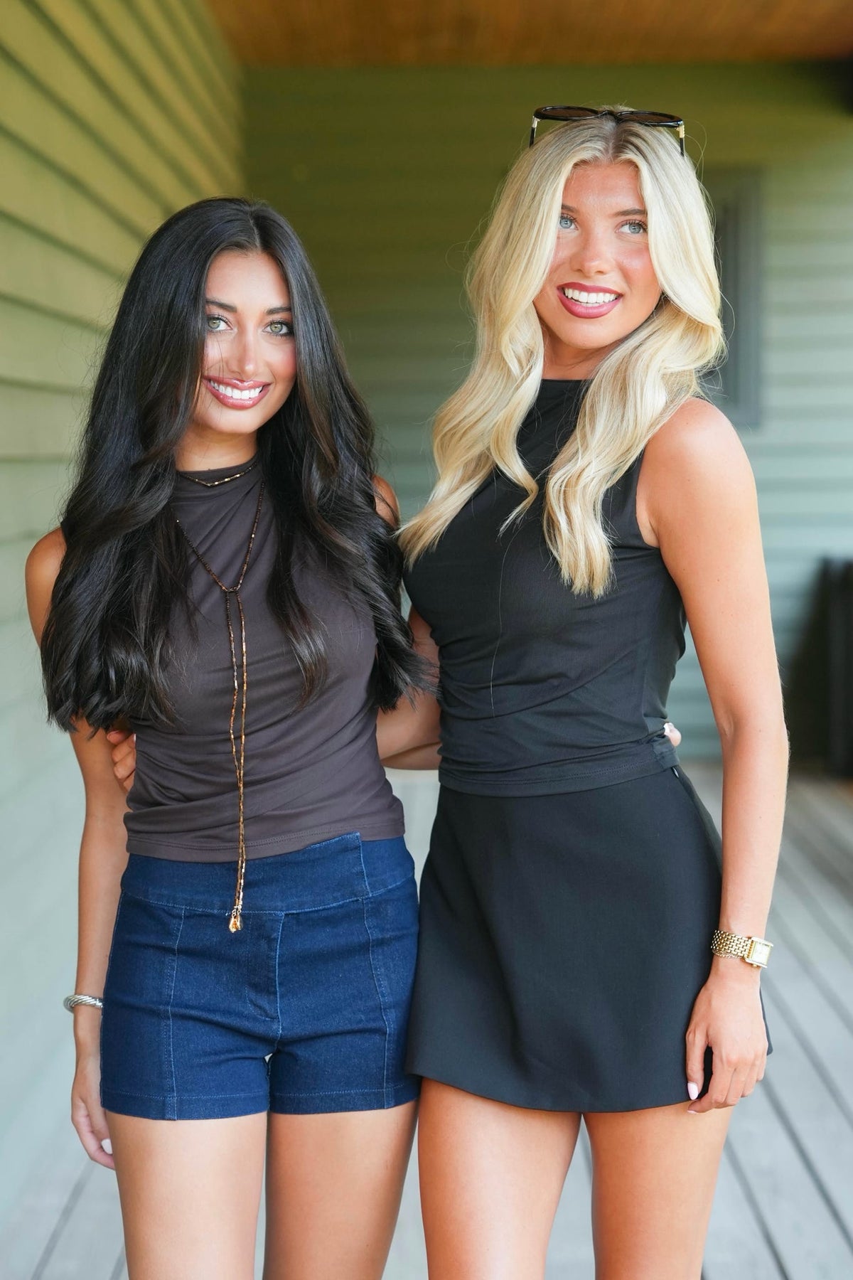 Two women standing together on a wooden porch. The woman on the left wears a dark brown sleeveless top, blue denim shorts, and brown knee-high boots.