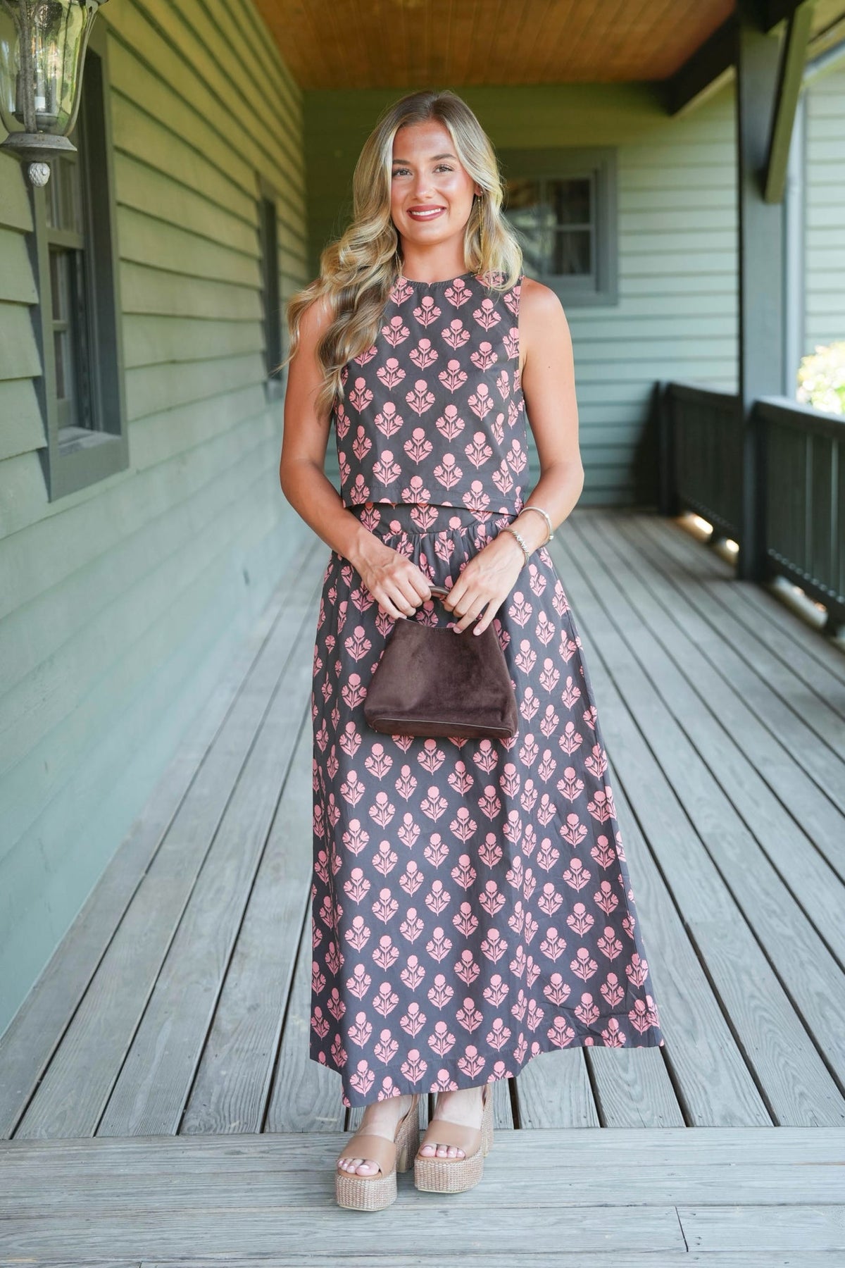 A woman stands on a wooden porch wearing a sleeveless midi dress with a pink and brown floral pattern, holding a brown handbag.