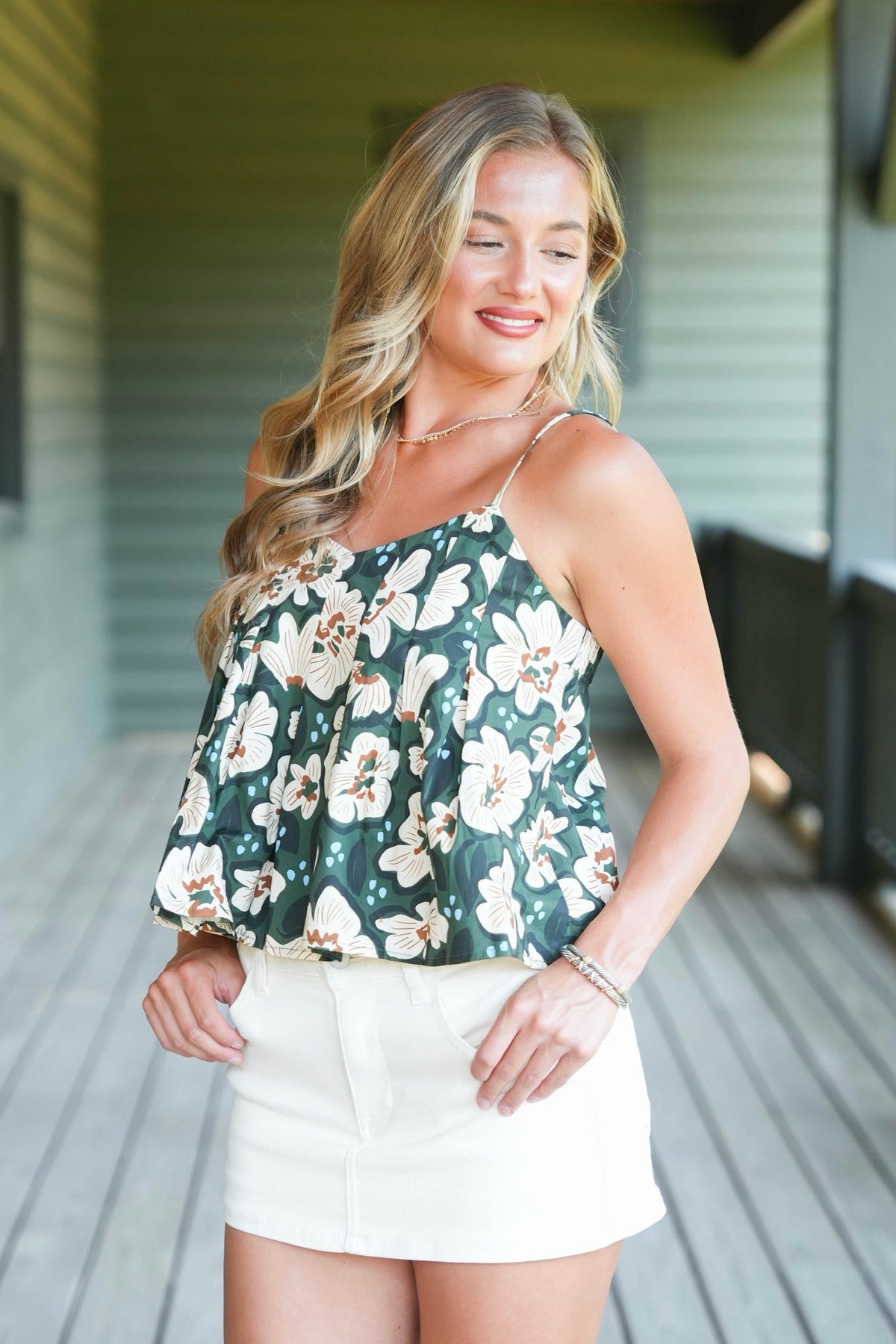 A woman modeling a dark green floral camisole top with white and beige flowers, paired with a white mini skirt, standing on a wooden porch.