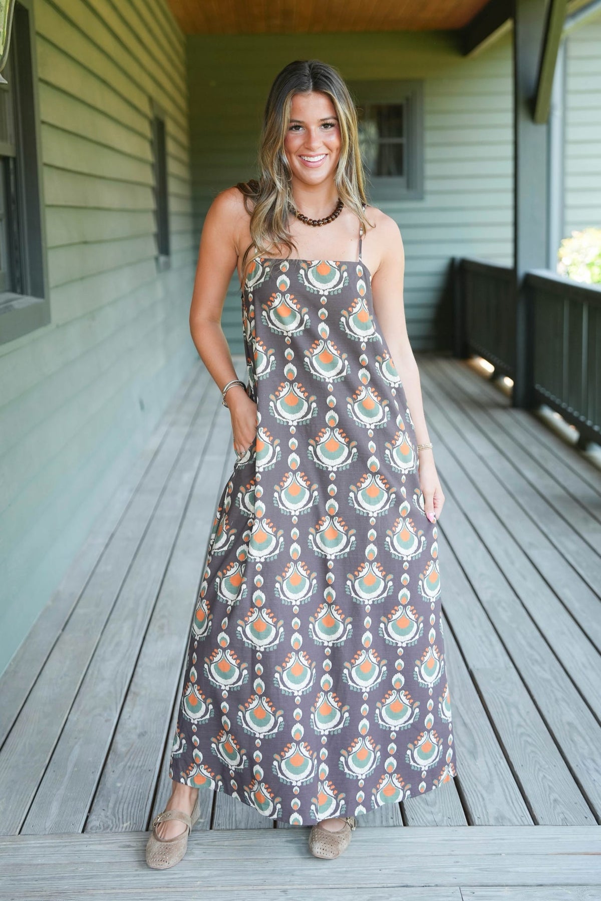 A woman wearing a sleeveless maxi dress with a brown, orange, and teal bohemian-style pattern, standing on a wooden deck in front of a house with green siding.