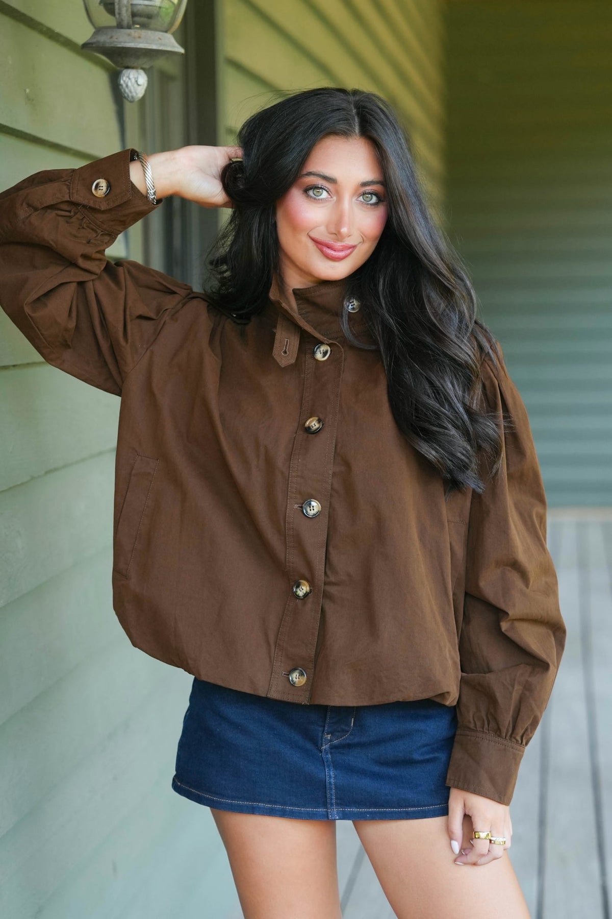 A woman wearing a brown button-up top and a blue denim mini skirt, posing on a porch with a green siding wall in the background.