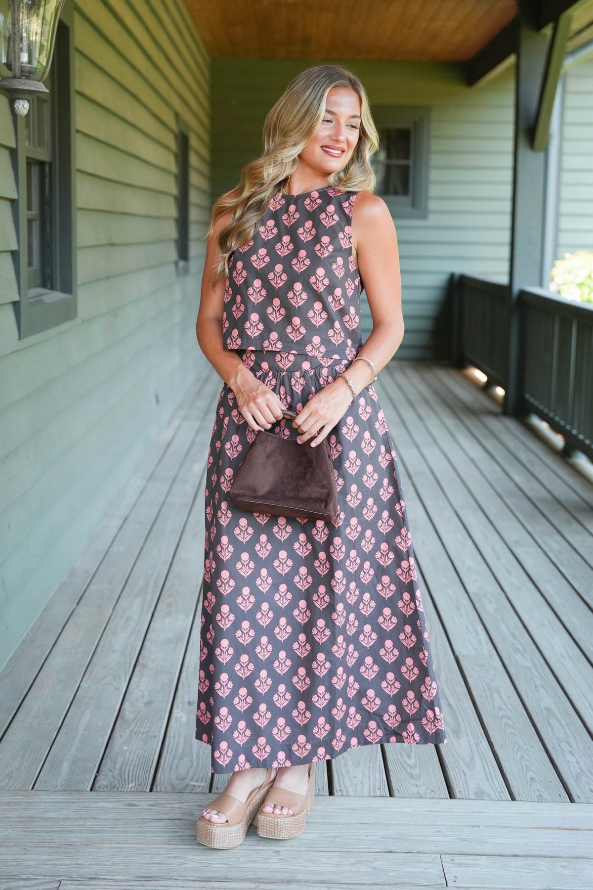 A woman stands on a wooden porch wearing a sleeveless, high-neck maxi dress with a brown base and a pink floral pattern. She is holding a small brown handbag and wearing beige platform sandals.