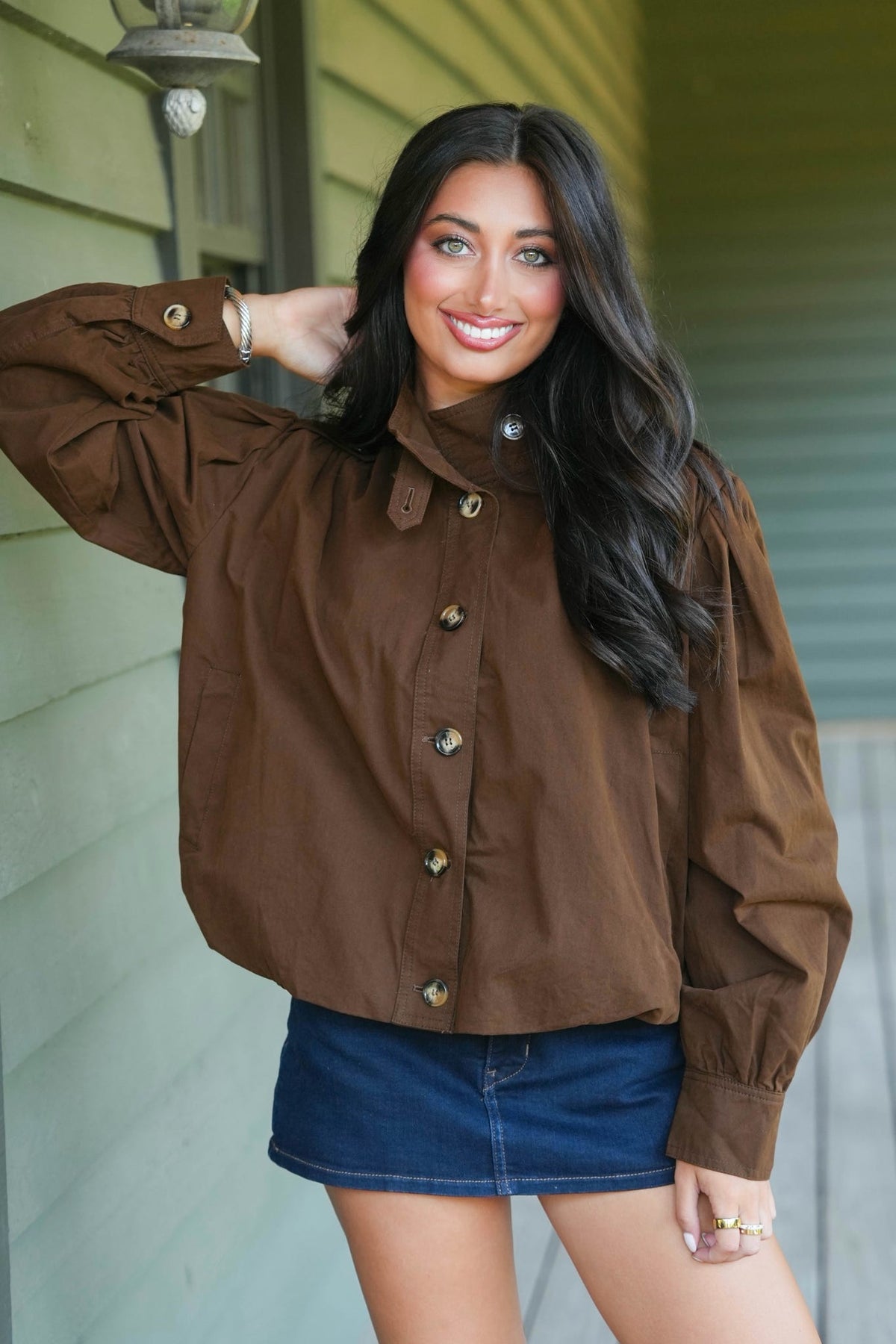 A woman wearing a brown button-up top and a blue denim mini skirt, posing on a porch with a green siding wall in the background.