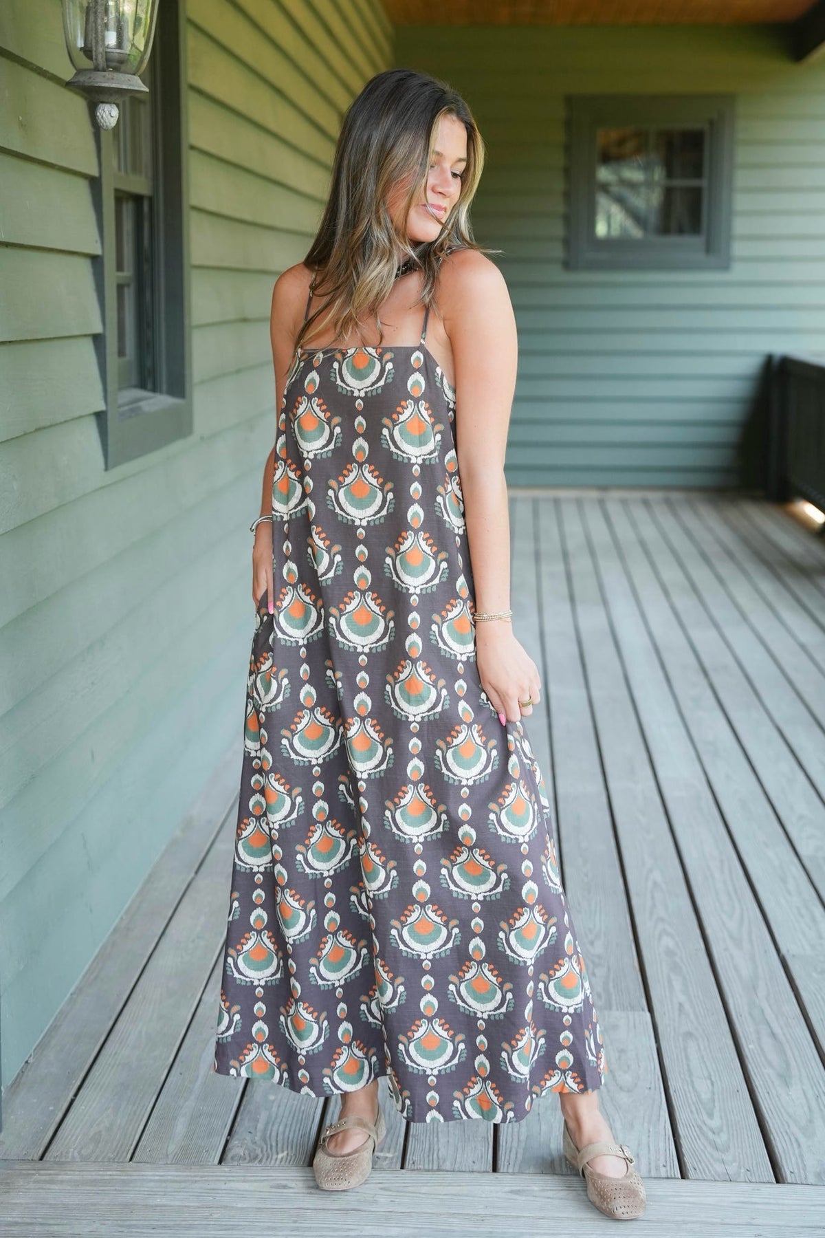A woman wearing a sleeveless maxi dress with a brown, orange, and teal bohemian-style pattern, standing on a wooden deck in front of a house with green siding.