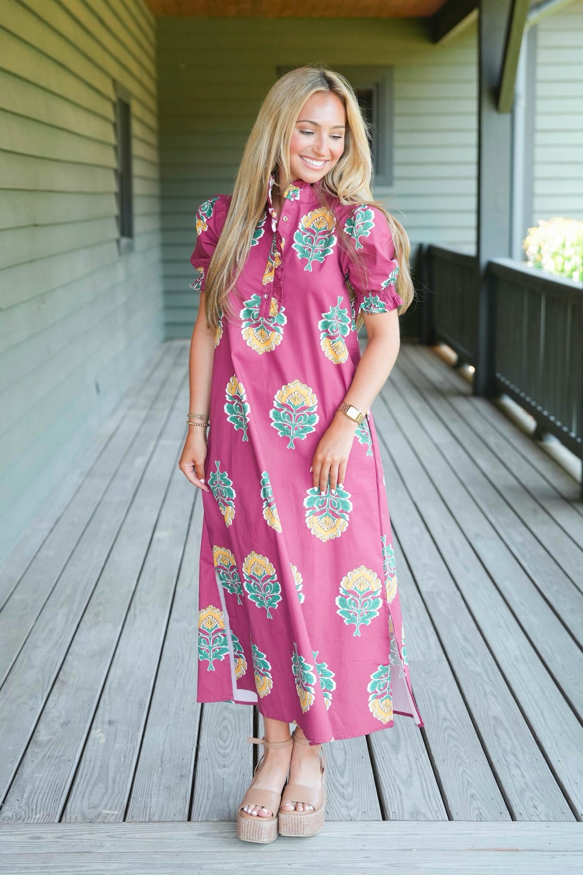A woman modeling a pink midi dress with a colorful floral print, standing on a wooden porch.