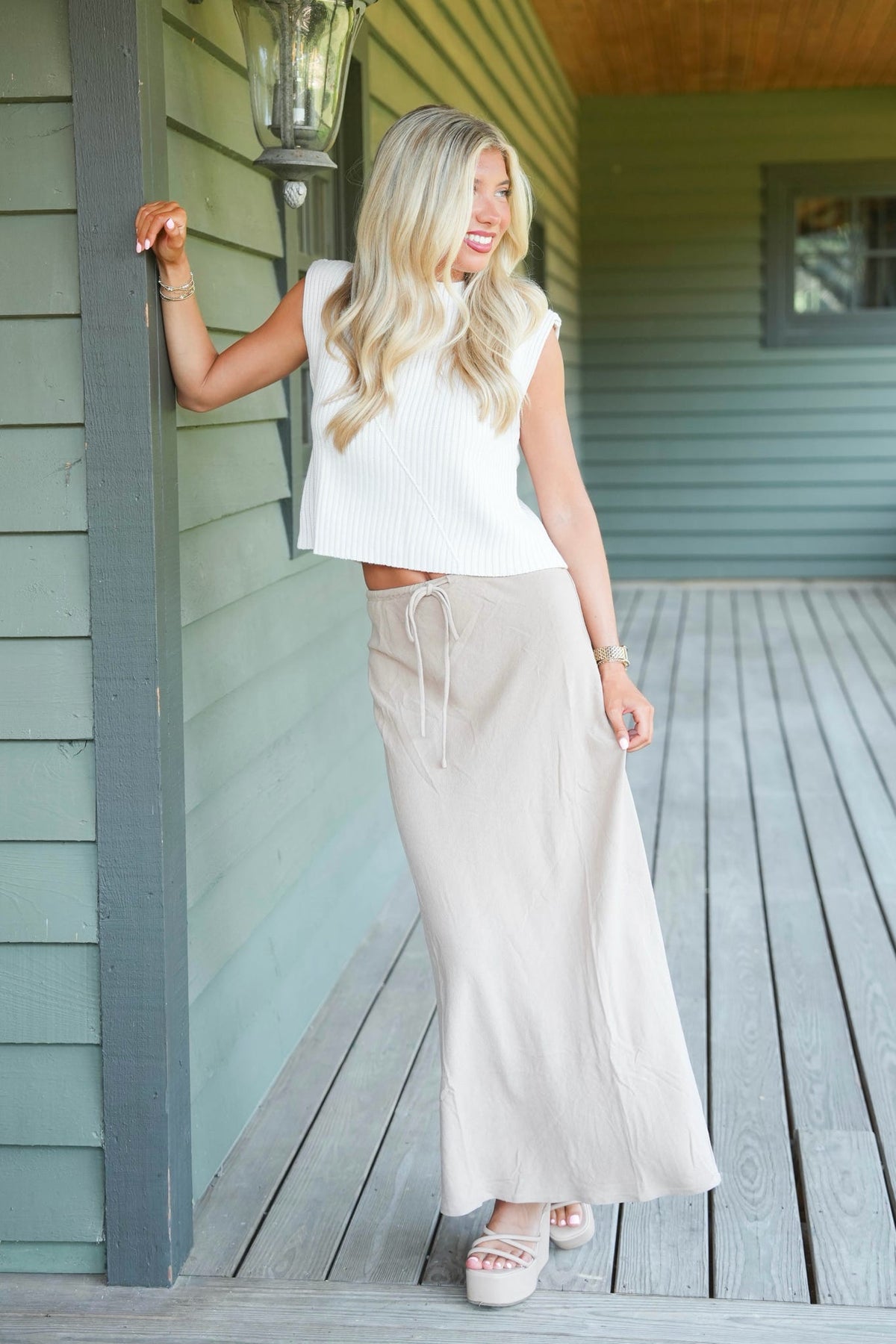 A smiling blonde woman is posing wearing a cream knitted sweater tank top paired with a beige maxi skirt. Standing, leaned in front of a green porch.