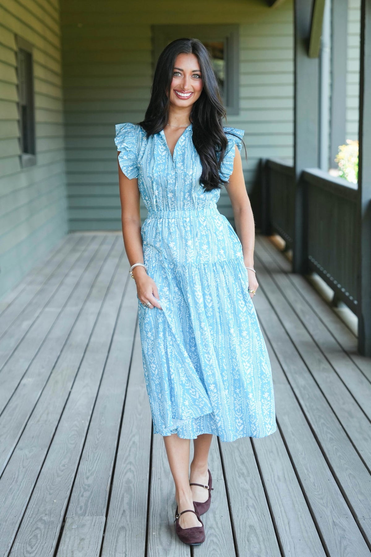 A woman stands on a wooden porch wearing a light blue midi dress with a white floral pattern, ruffled cap sleeves, and a V-neckline.