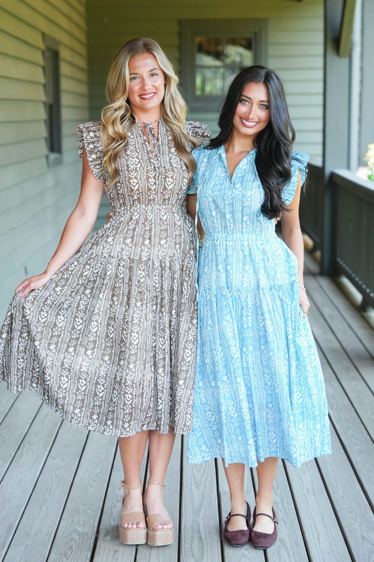 Two women modeling midi dresses on a wooden porch. The woman on the left wears a champagne beige dress with a black floral pattern and fringe details, while the woman on the right wears a light blue dress with a white floral pattern.