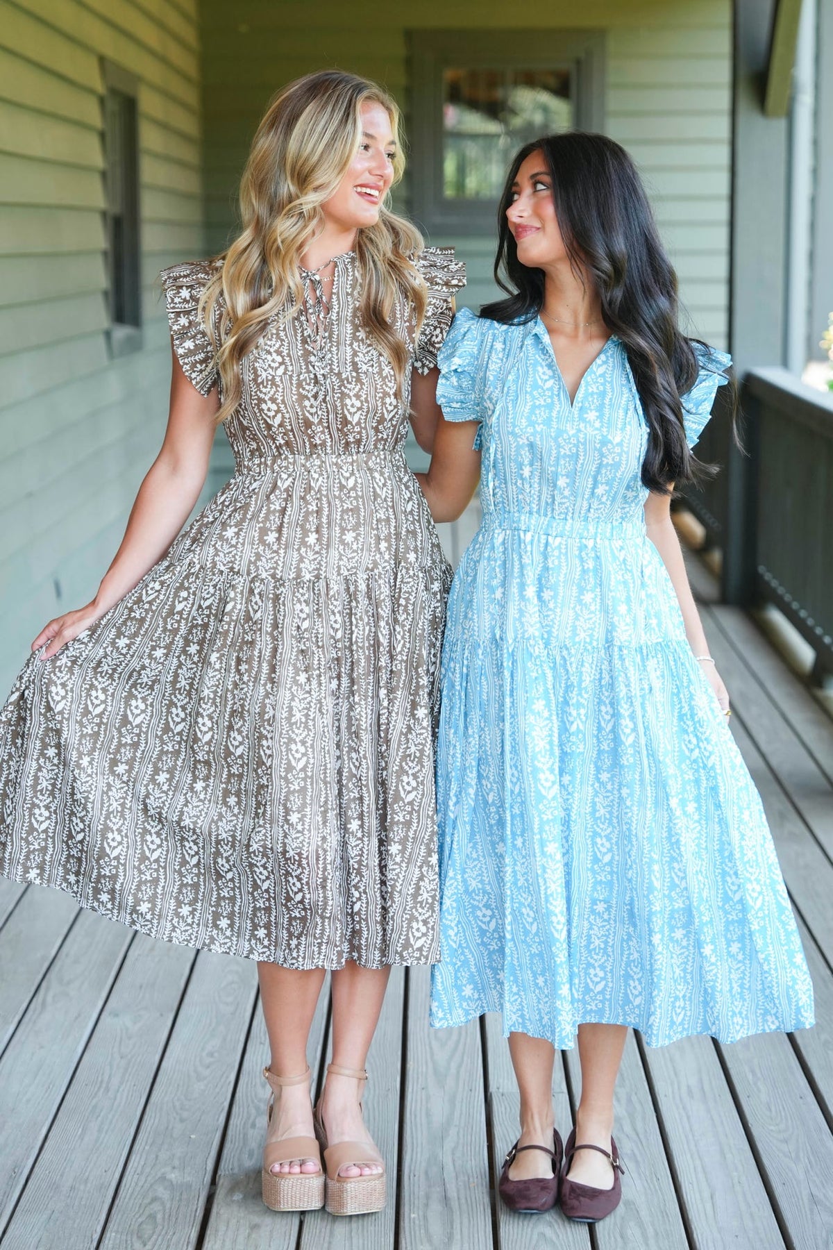Two women standing on a wooden porch wearing matching midi dresses with ruffled sleeves. The woman on the left wears a champagne beige dress with a black floral pattern, and the woman on the right wears a light blue dress with a white floral pattern.