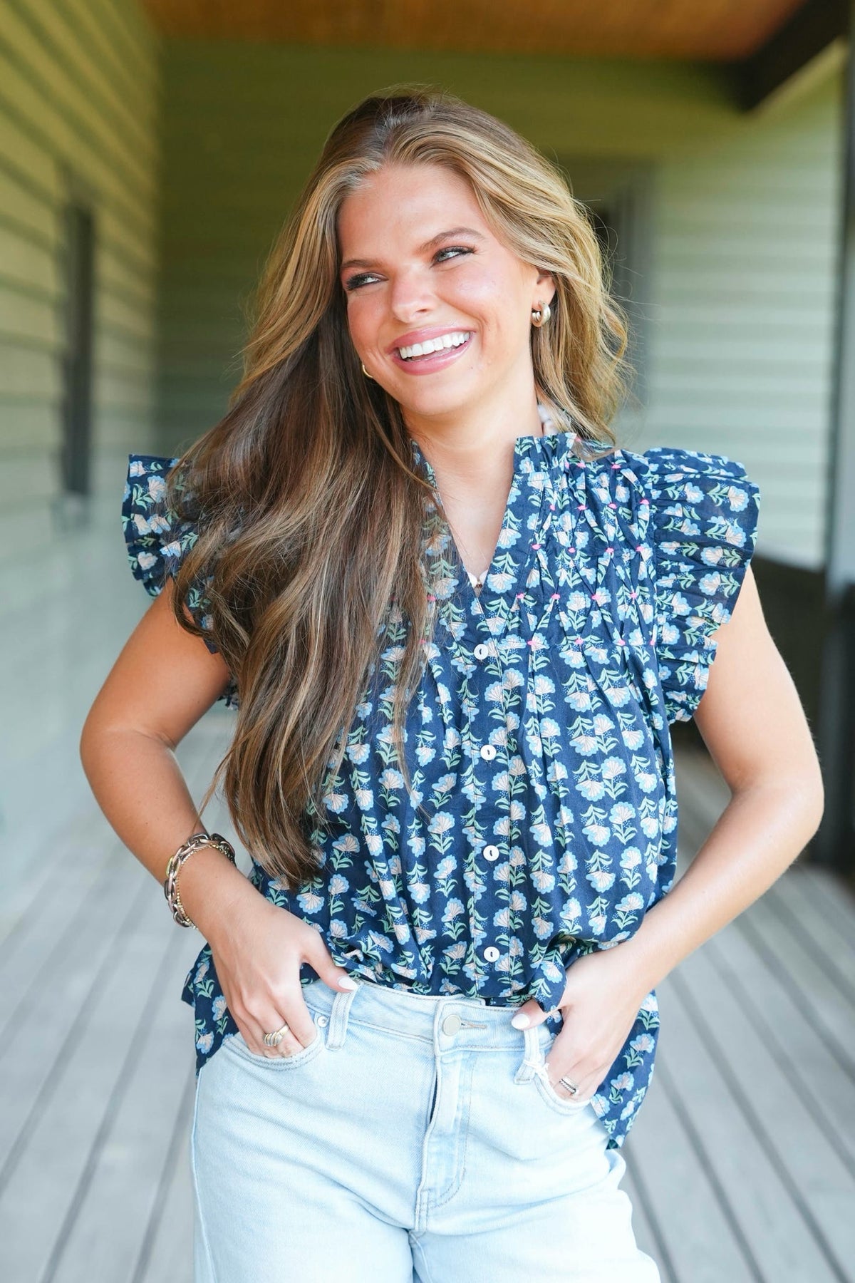 A smiling brunette woman wearing a navy sleeveless blouse with a green and pink floral pattern and ruffled shoulders, paired with white jeans, standing on a wooden porch.