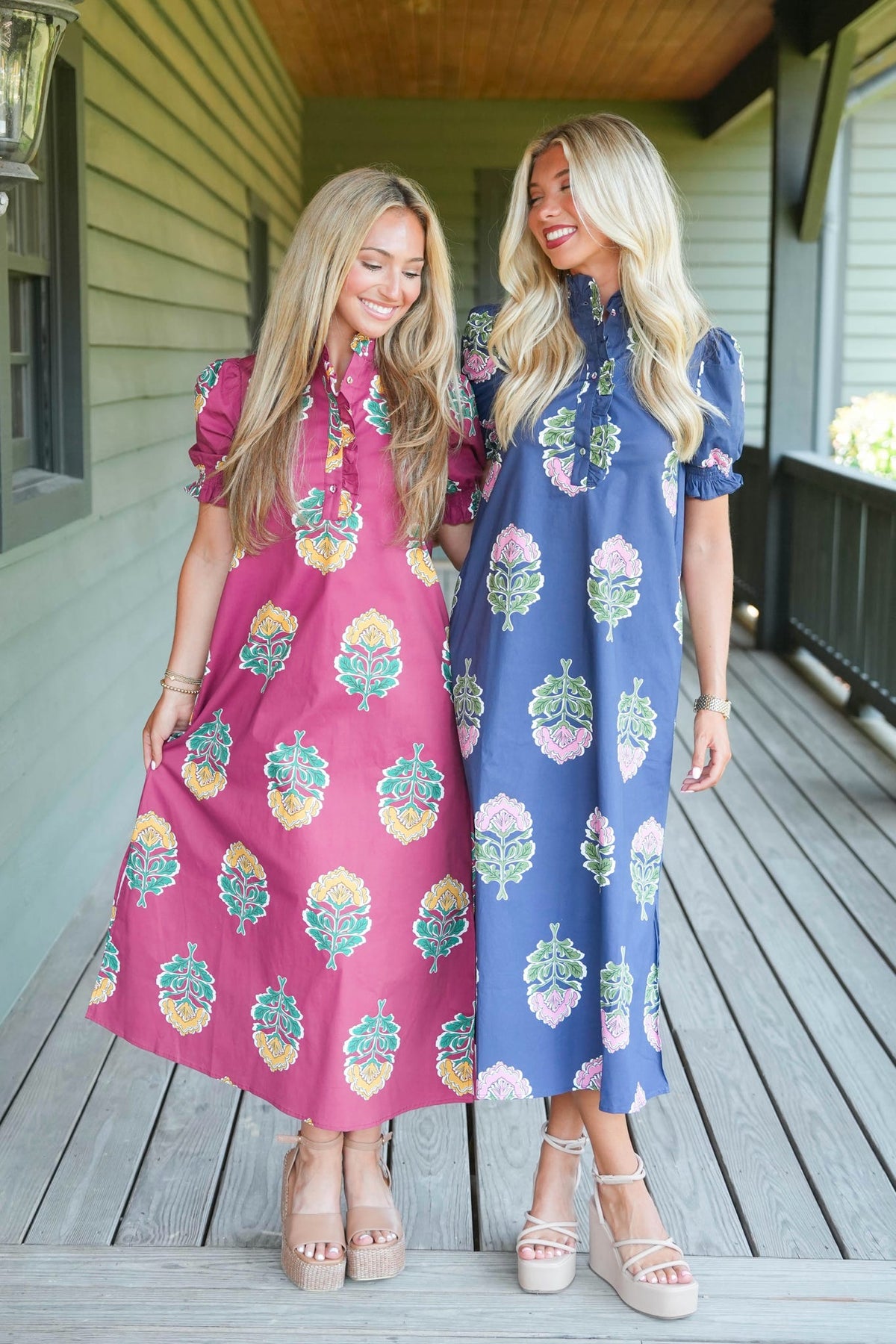 Two women modeling matching short-sleeve midi dresses with a colorful floral block print, standing on a wooden porch. The dress on the left is magenta pink, and the dress on the right is blue.