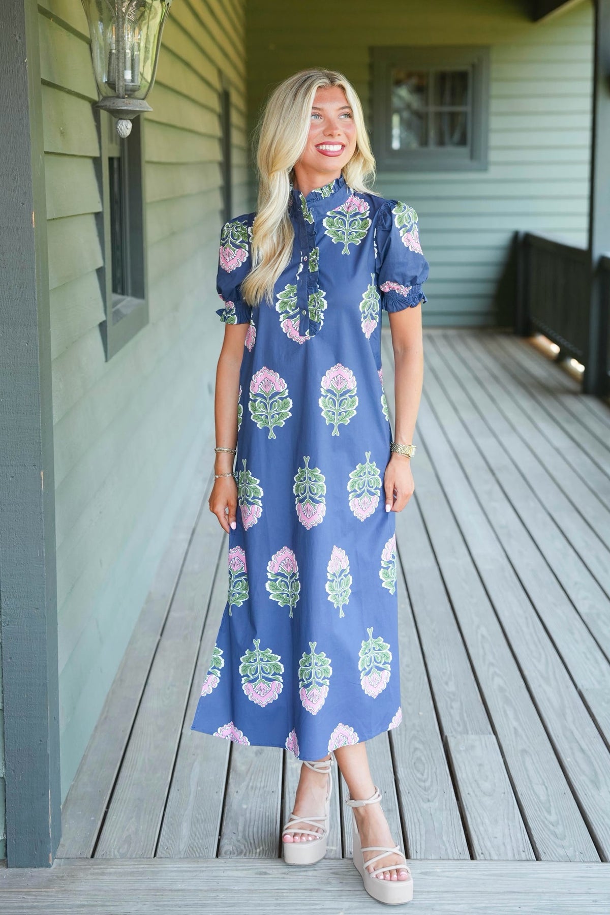 A woman stands on a wooden porch wearing a blue midi dress with a pink and green floral pattern, paired with beige platform sandals.