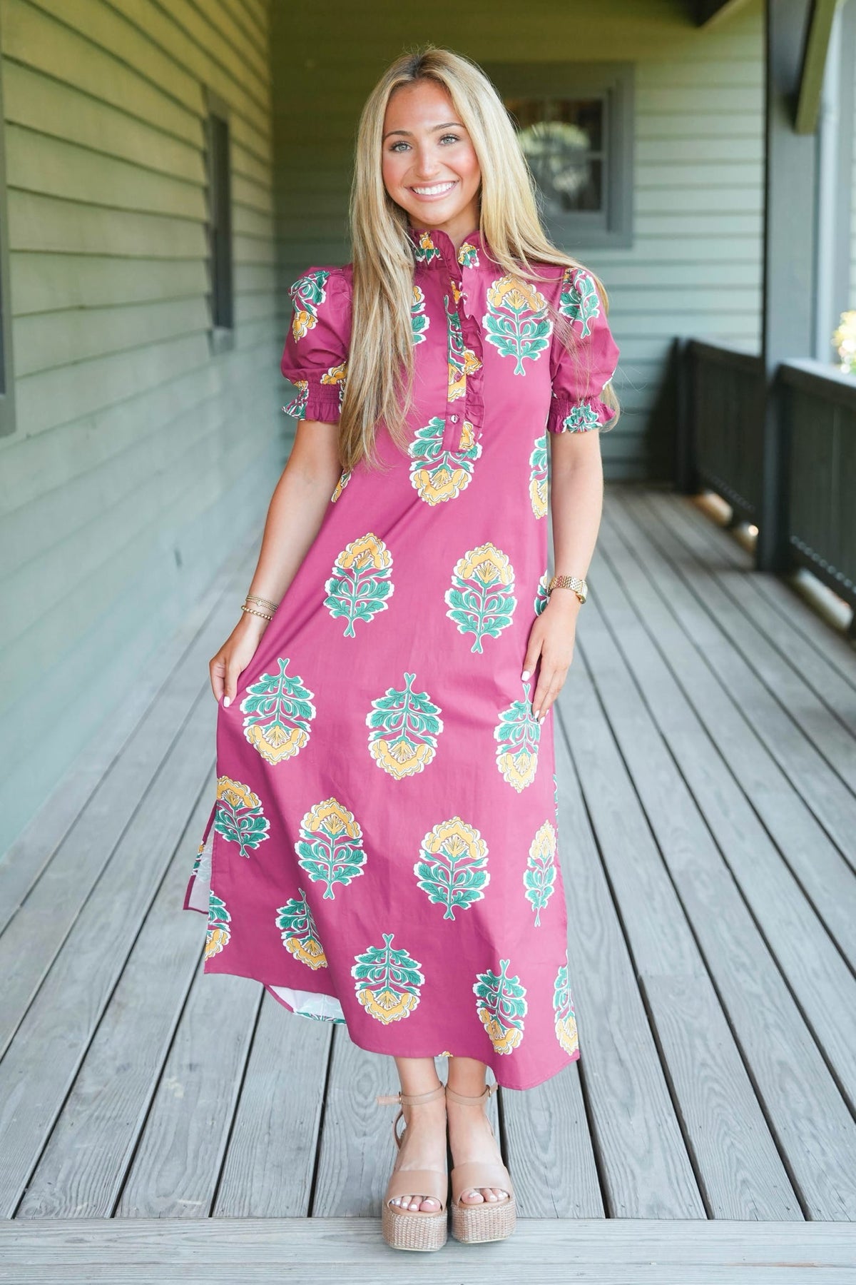 A woman stands on a wooden porch wearing a pink midi dress with a colorful floral pattern, puff sleeves, and a tie-neck detail.