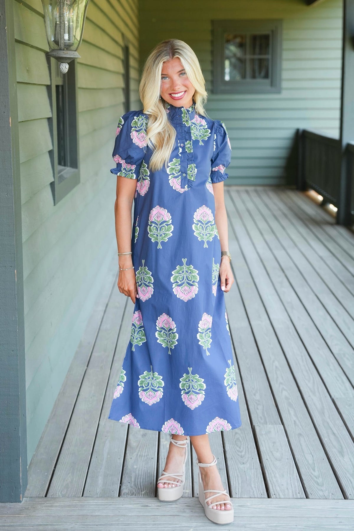 A woman stands on a wooden porch wearing a blue midi dress with a pink and green floral pattern, paired with beige platform sandals.