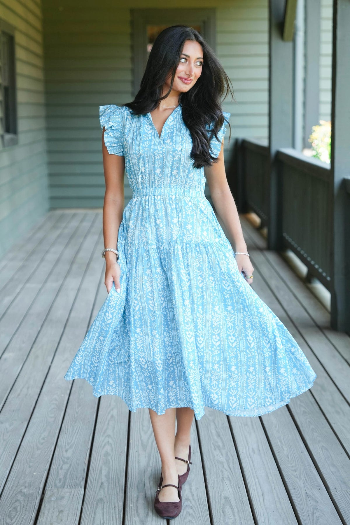 A woman wearing a light blue lace midi dress with ruffled sleeves and a V-neckline, walking on a wooden porch.