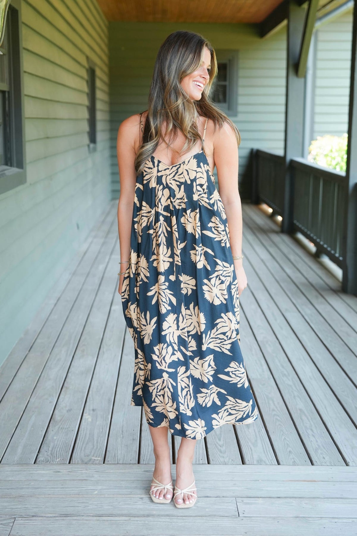 A woman wearing a black midi dress with a beige tropical leaf print stands on a wooden porch.