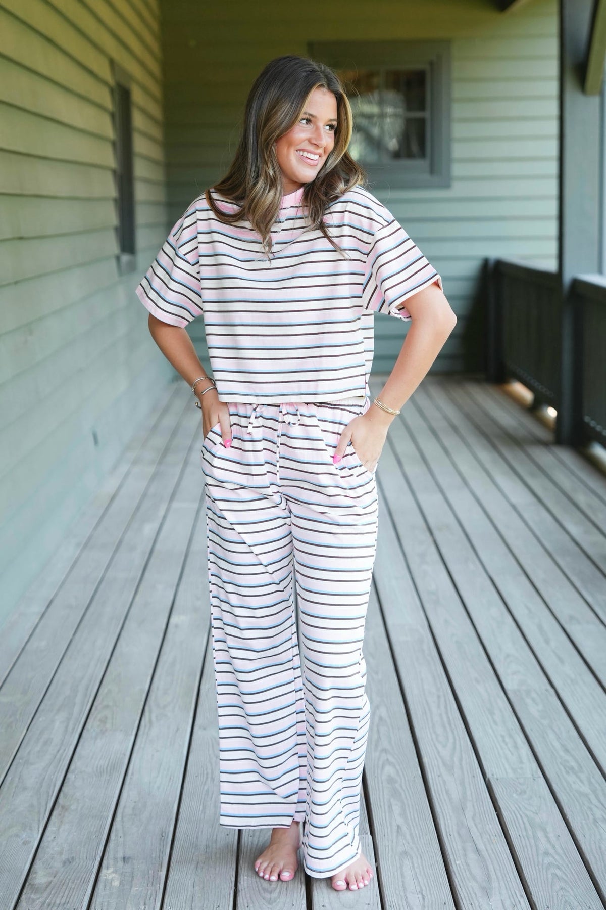 A woman stands barefoot on a wooden porch wearing a matching short-sleeve striped pajama top and wide-leg pajama pants in white with thin navy and pink horizontal stripes.