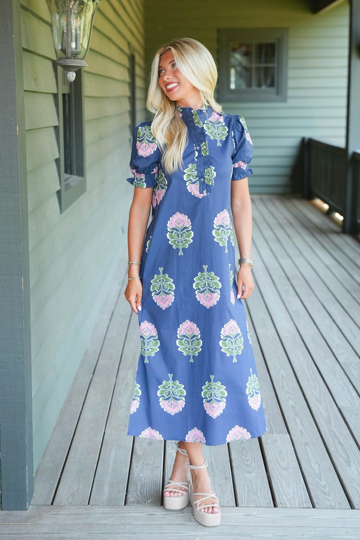 A woman stands on a wooden porch wearing a blue midi dress with a pink and green floral pattern, paired with beige platform sandals.