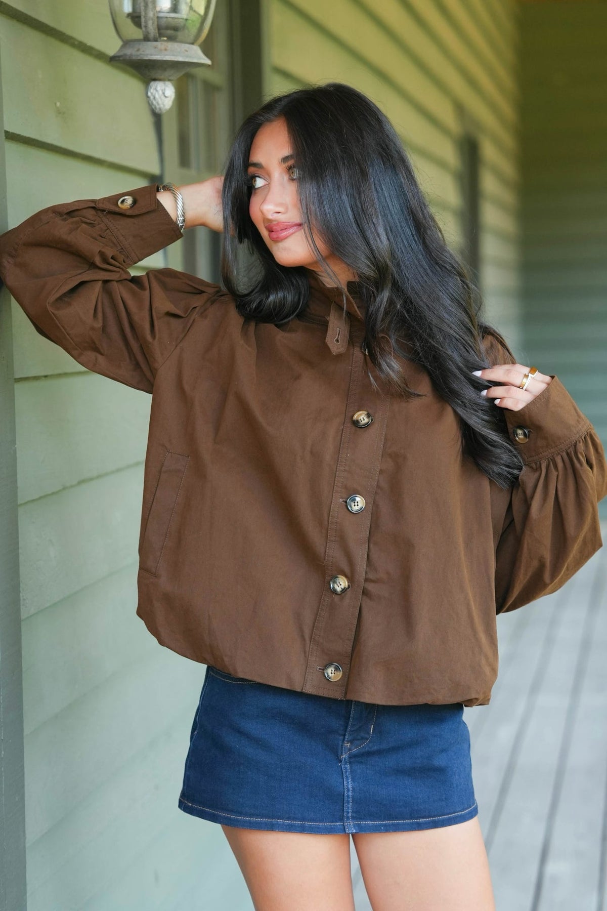 A woman wearing a brown button-up shirt with long puff sleeves and a blue denim mini skirt, standing on a porch in front of a light green house wall.