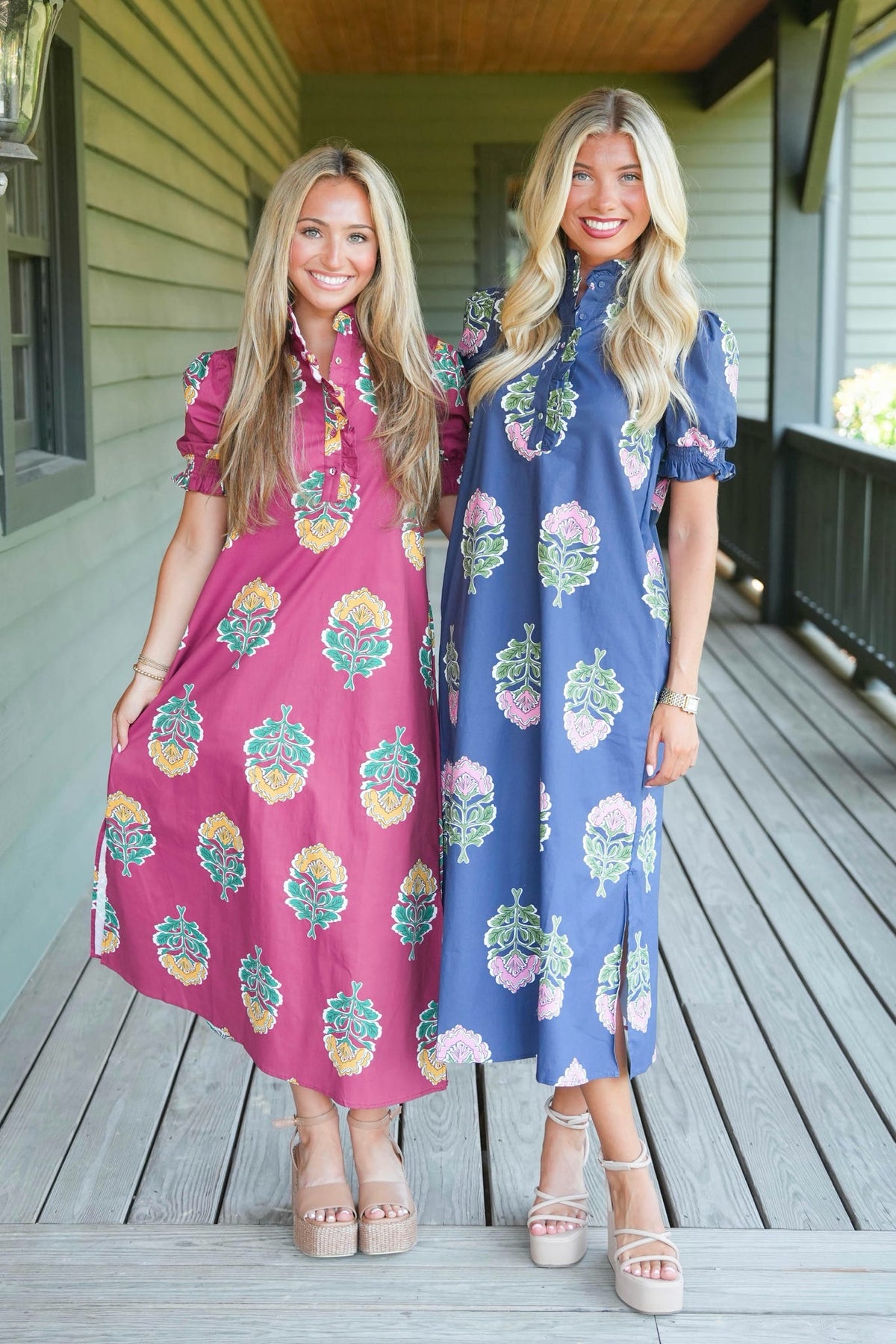 Two women modeling short puff-sleeve midi dresses with colorful floral prints, standing on a wooden porch. One dress is pink with yellow, green, and blue motifs, and the other is blue with pink, green, and yellow motifs.