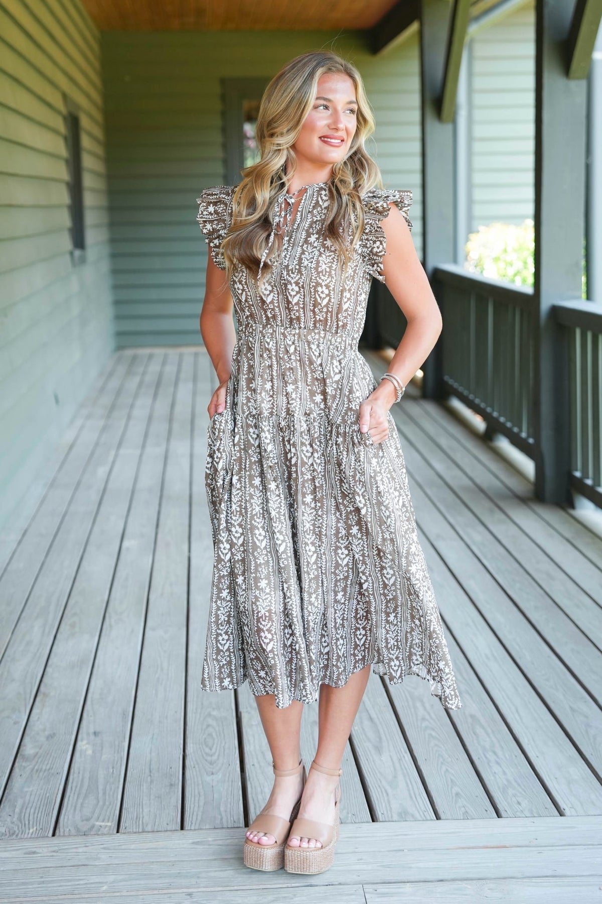 A woman stands on a wooden porch wearing a sleeveless, midi-length lace dress with ruffled cap sleeves and a silver and beige floral pattern. She is wearing beige platform sandals.