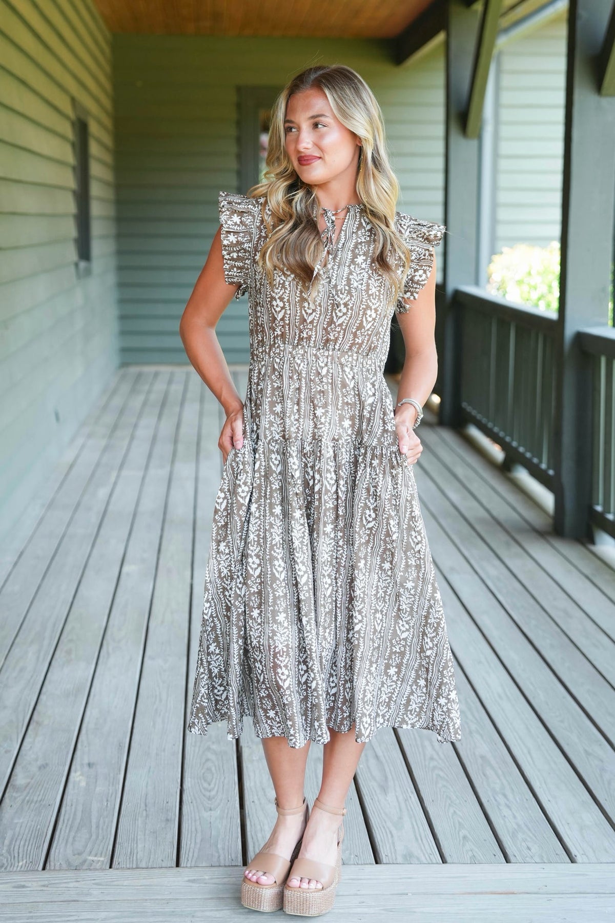 A woman stands on a wooden porch wearing a sleeveless, midi-length lace dress with ruffled cap sleeves and a silver and beige floral pattern. She is wearing beige platform sandals.