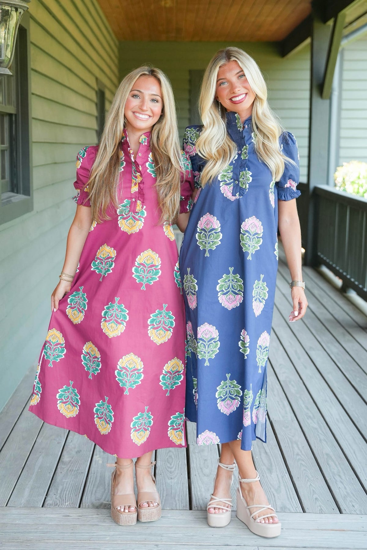 Two women modeling short puff-sleeve midi dresses with colorful floral prints, standing on a wooden porch. One dress is pink with yellow, green, and blue motifs, and the other is blue with pink, green, and yellow motifs.