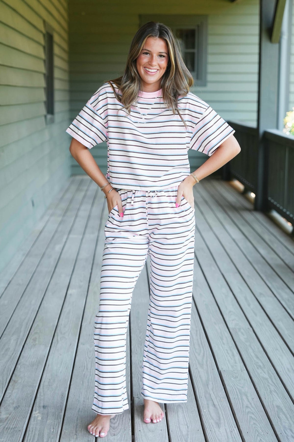A woman stands barefoot on a wooden porch wearing a matching short-sleeve striped pajama top and wide-leg pajama pants in white with thin navy and pink horizontal stripes.