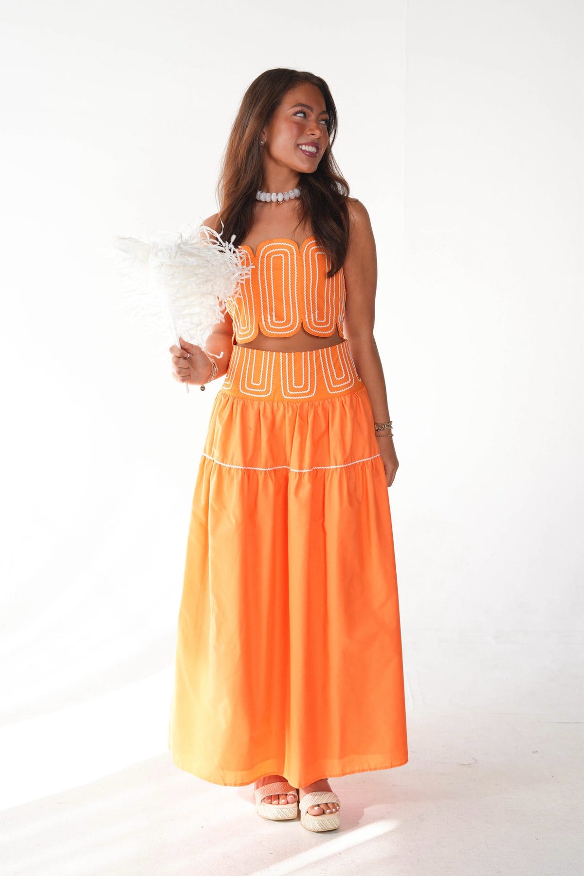 A woman modeling a bright orange two-piece outfit consisting of a sleeveless crop top and a matching tiered maxi skirt, with white geometric embroidered patterns. She is holding a white feather fan against a plain white studio background.
