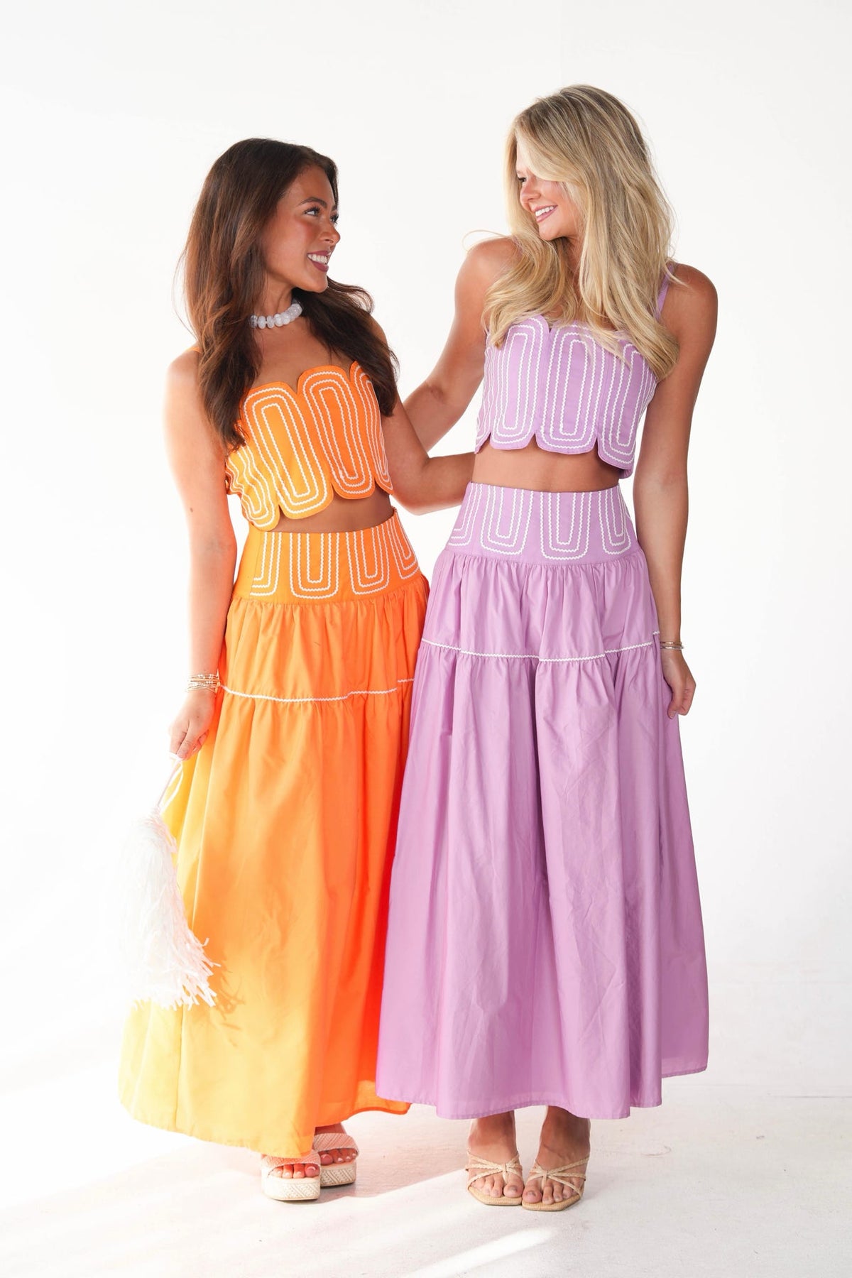 Two women modeling matching two-piece outfits against a white studio background. The woman on the left wears an orange and yellow set, and the woman on the right wears a lavender and light purple set.