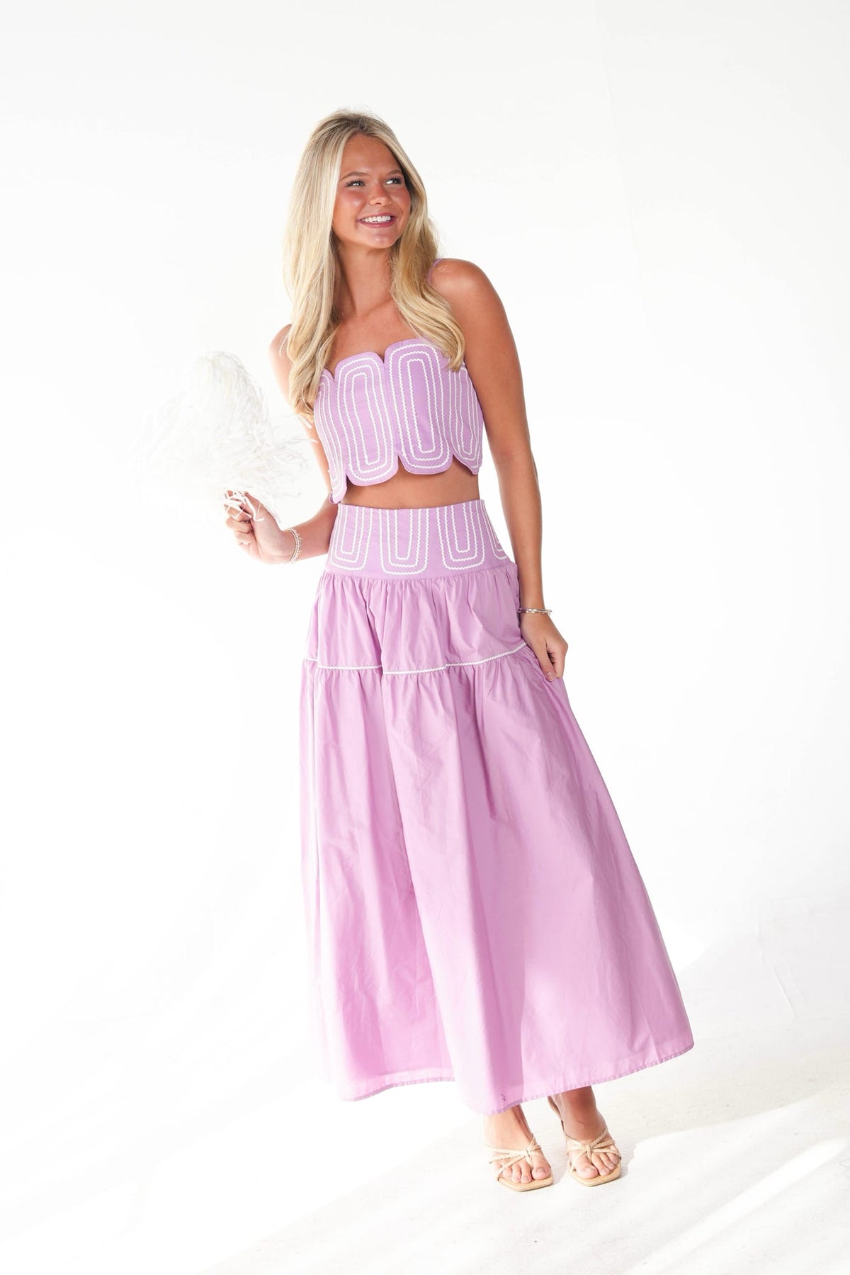 A woman modeling a lavender and white two-piece set consisting of a strapless crop top and a matching tiered maxi skirt, holding a white textured clutch against a white studio background.