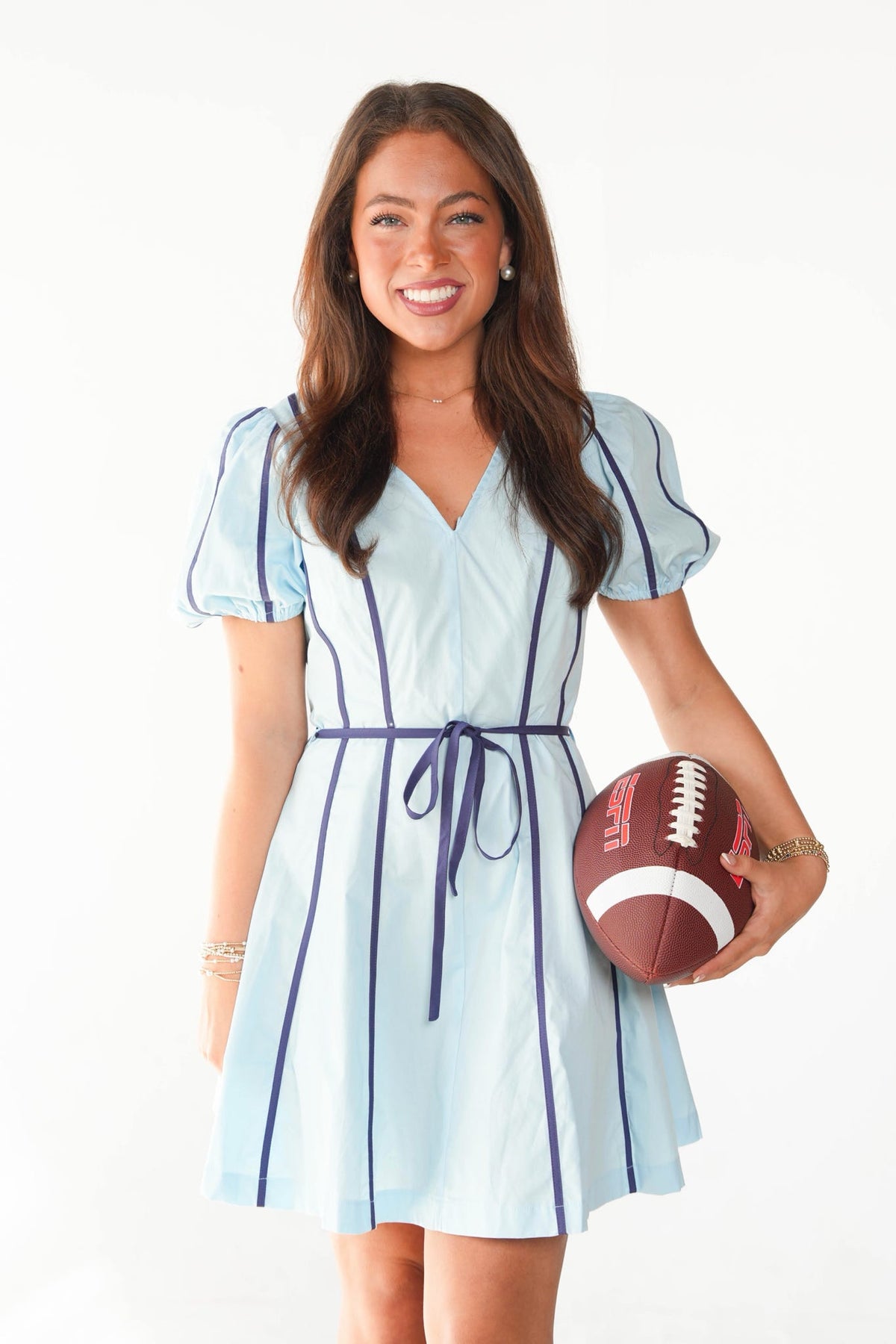 A woman modeling a light blue short-sleeve dress with dark navy vertical piping and a matching waist tie, holding a brown football against a plain white studio background.