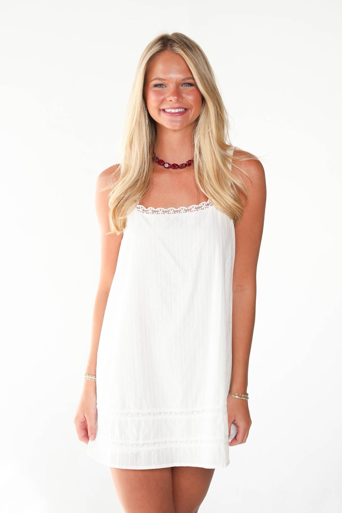A smiling blonde woman wearing a white sleeveless mini dress with delicate lace trim at the neckline, photographed against a plain white studio background.