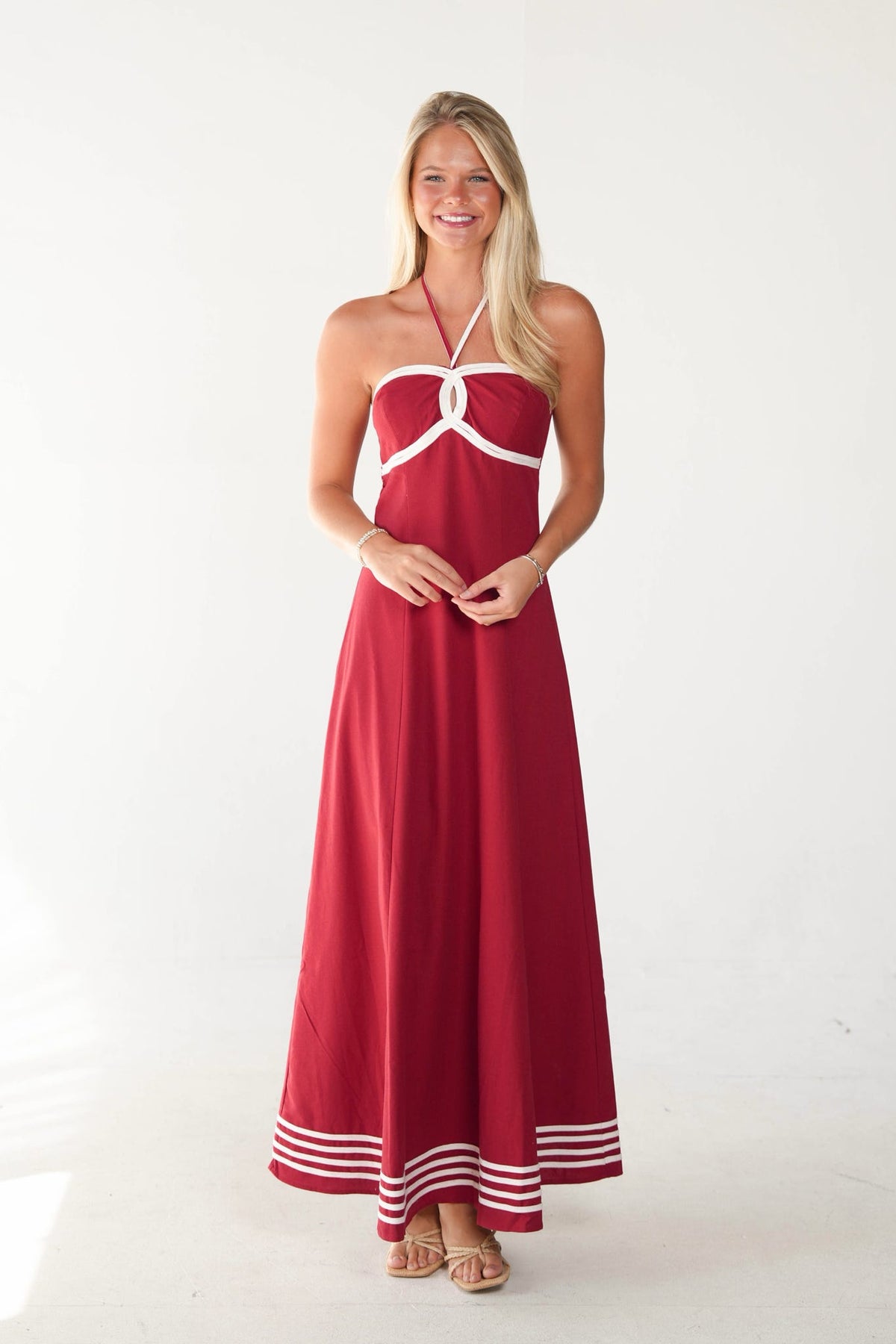 A woman modeling a red halter maxi dress with white contrast trim and striped detailing at the hem, standing against a plain white studio background.