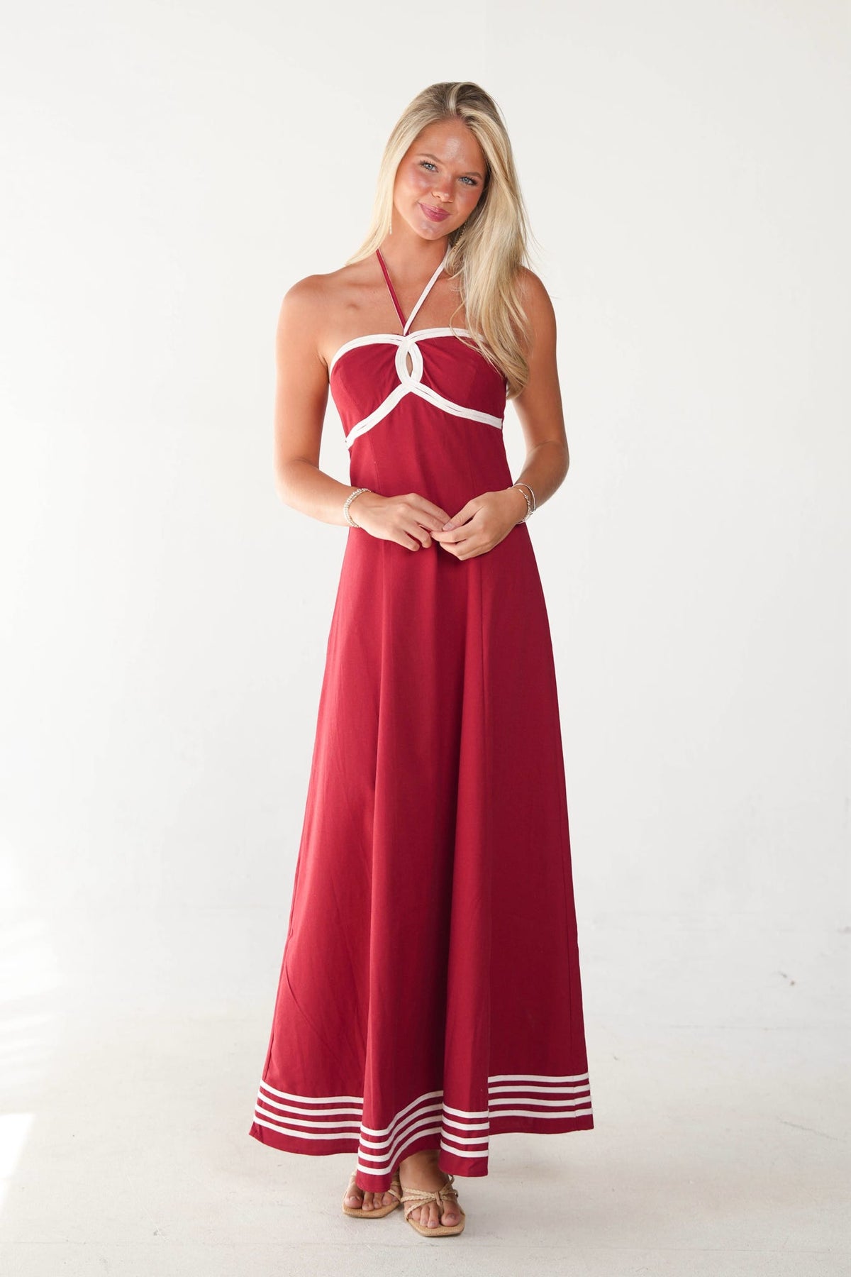 A woman modeling a red halter-neck maxi dress with white trim details, including a crossover at the bust and double stripes near the hem, set against a plain white studio background.