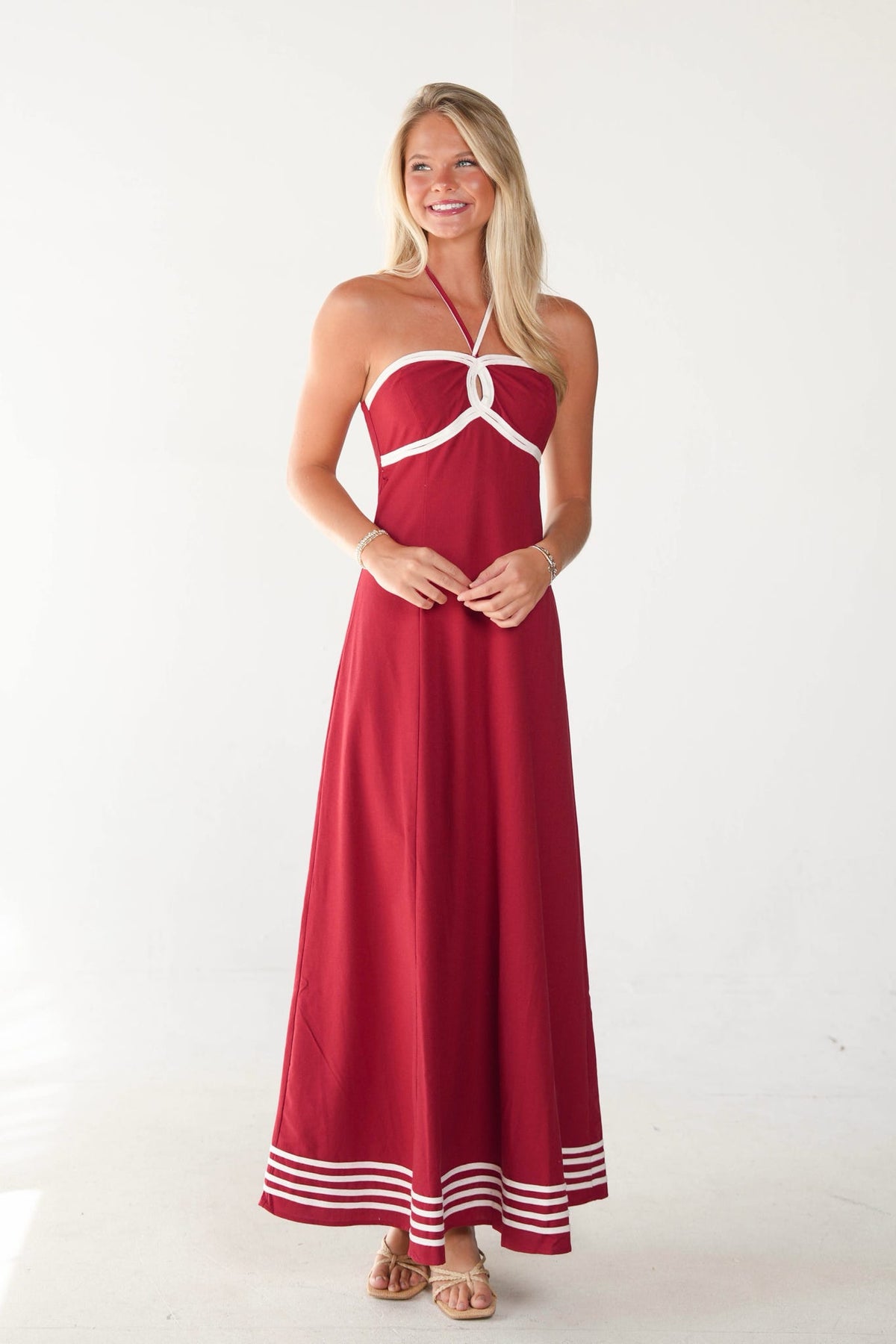 A woman modeling a red halter-neck maxi dress with white contrast trim at the bust and along the hem, standing against a plain white studio background.