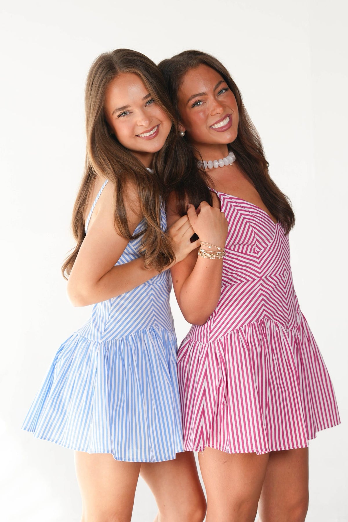 Two young women posing together against a white studio background, each wearing a short pleated dress with vertical stripes. The woman on the left wears a light blue and white striped dress, and the woman on the right wears a pink and white striped dress.