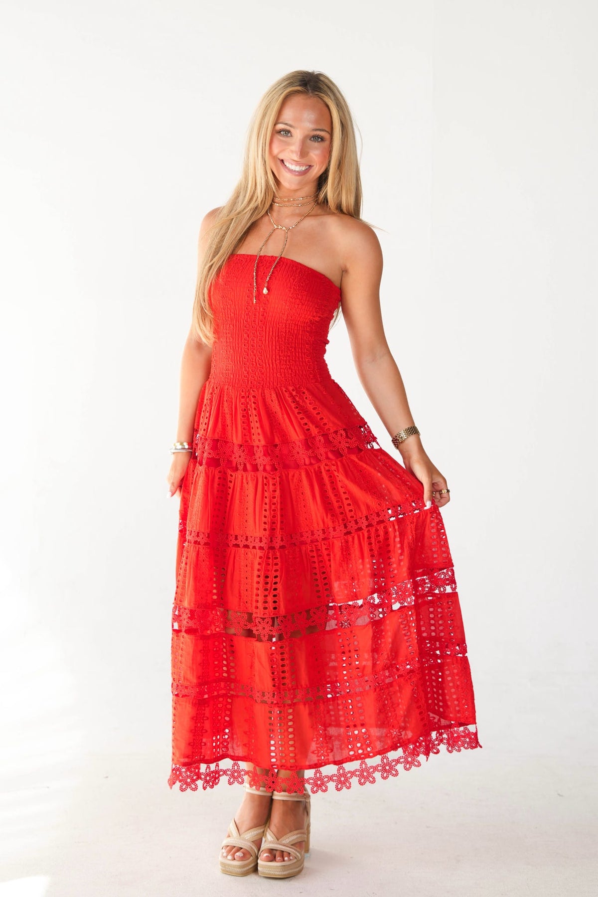 A woman modeling a bright red strapless maxi dress with intricate floral embroidery and tiered ruffles, standing against a plain white studio background.